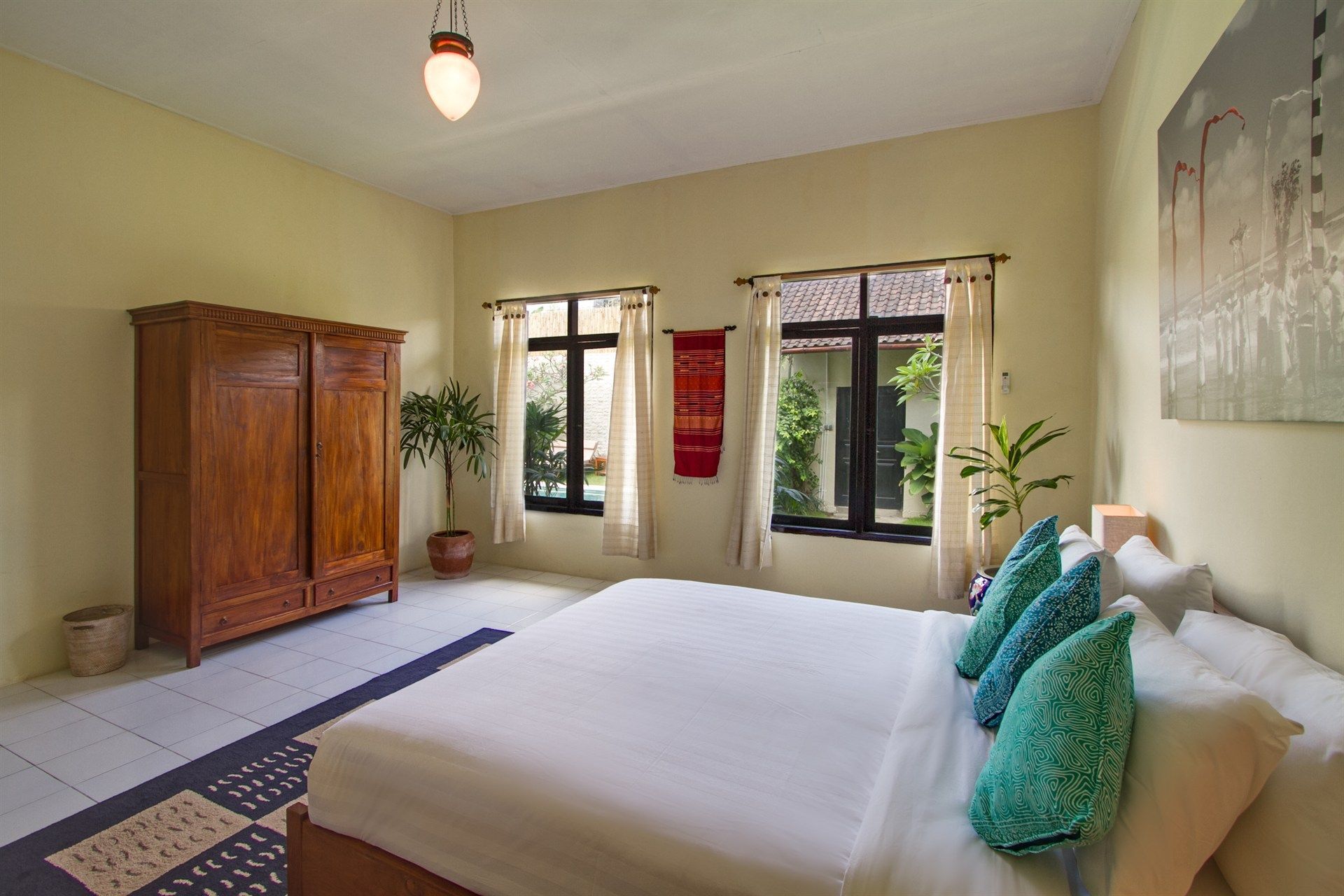 Bedroom with wooden wardrobe, bed with teal pillows, two windows overlooking greenery.