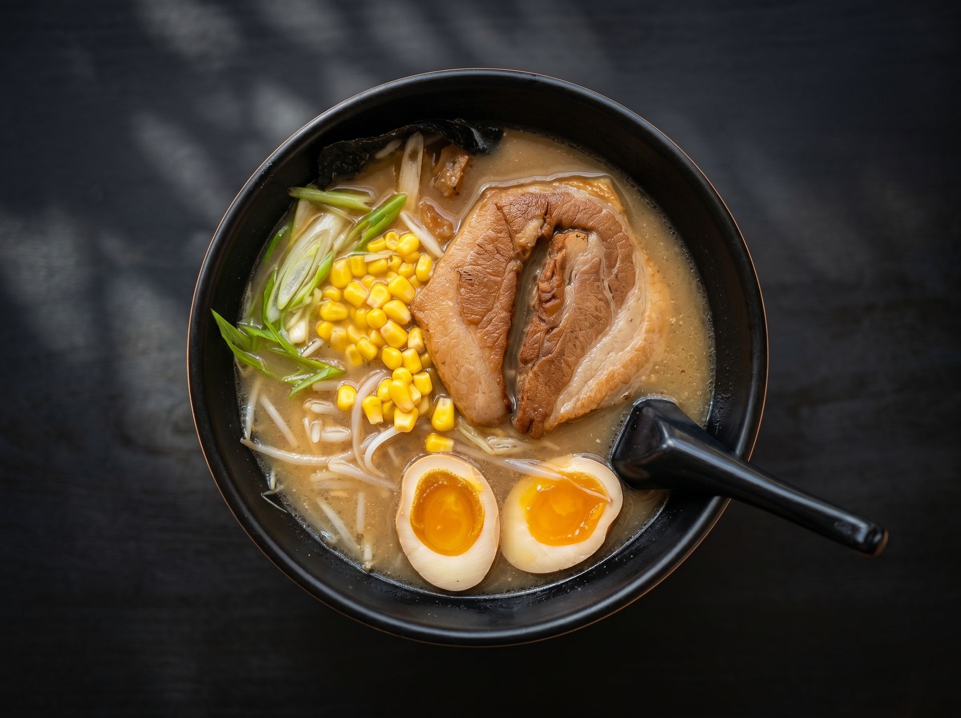 Black bowl of ramen with pork, corn, egg halves, and green onions on a dark table