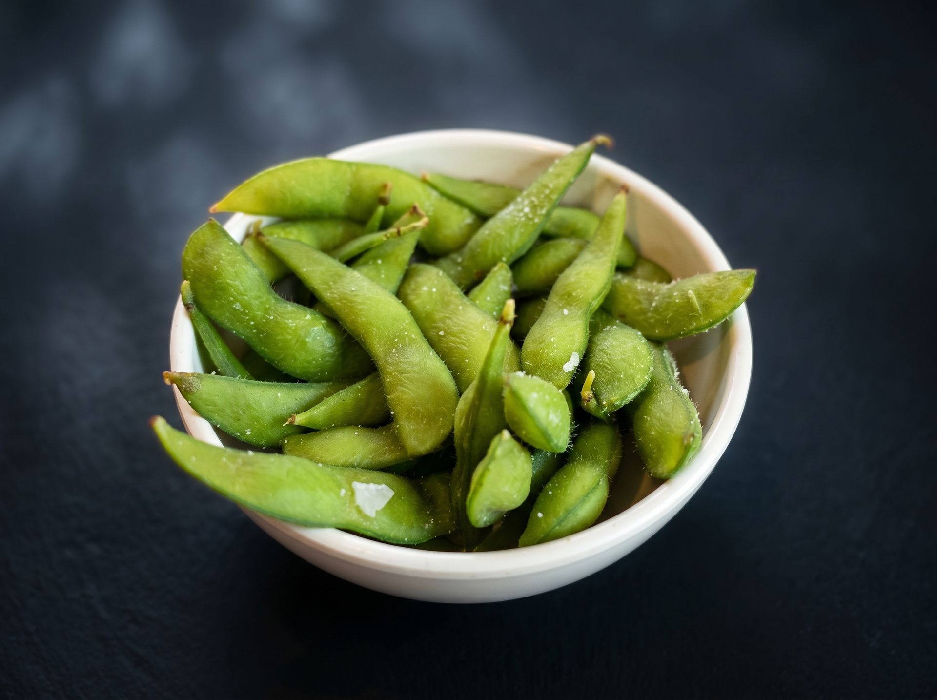 White bowl of green edamame pods on a dark surface