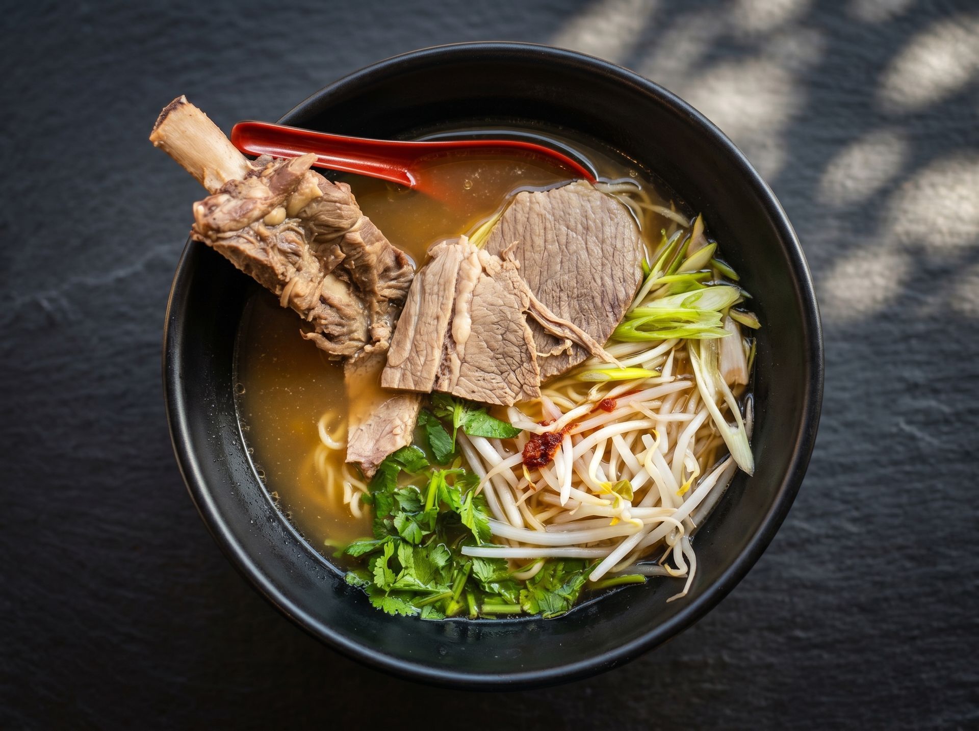 Bowl of beef noodle soup with greens and bean sprouts, topped with a large bone, on a dark table