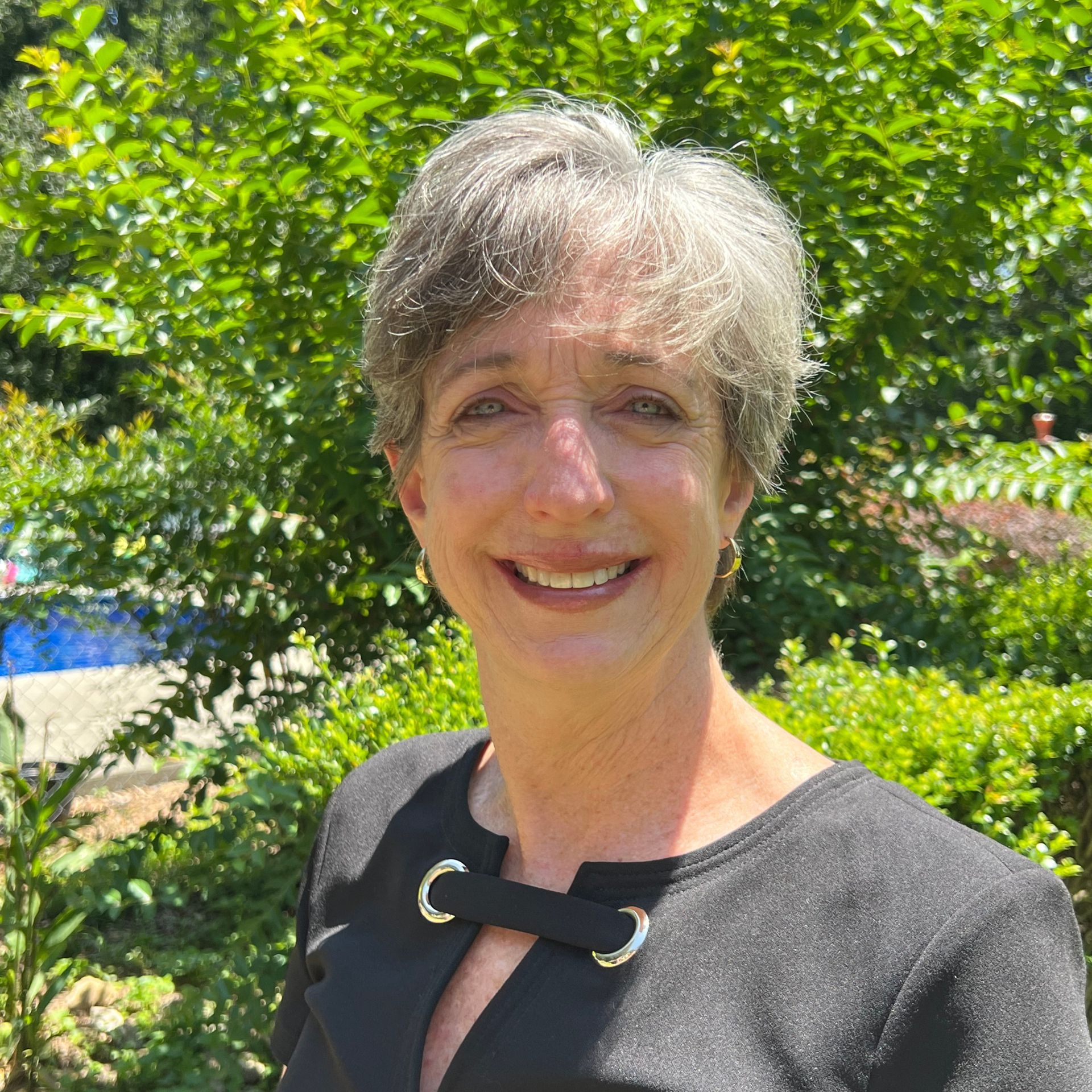 Woman with short gray hair smiles in front of green foliage, wearing a black top with silver hardware.