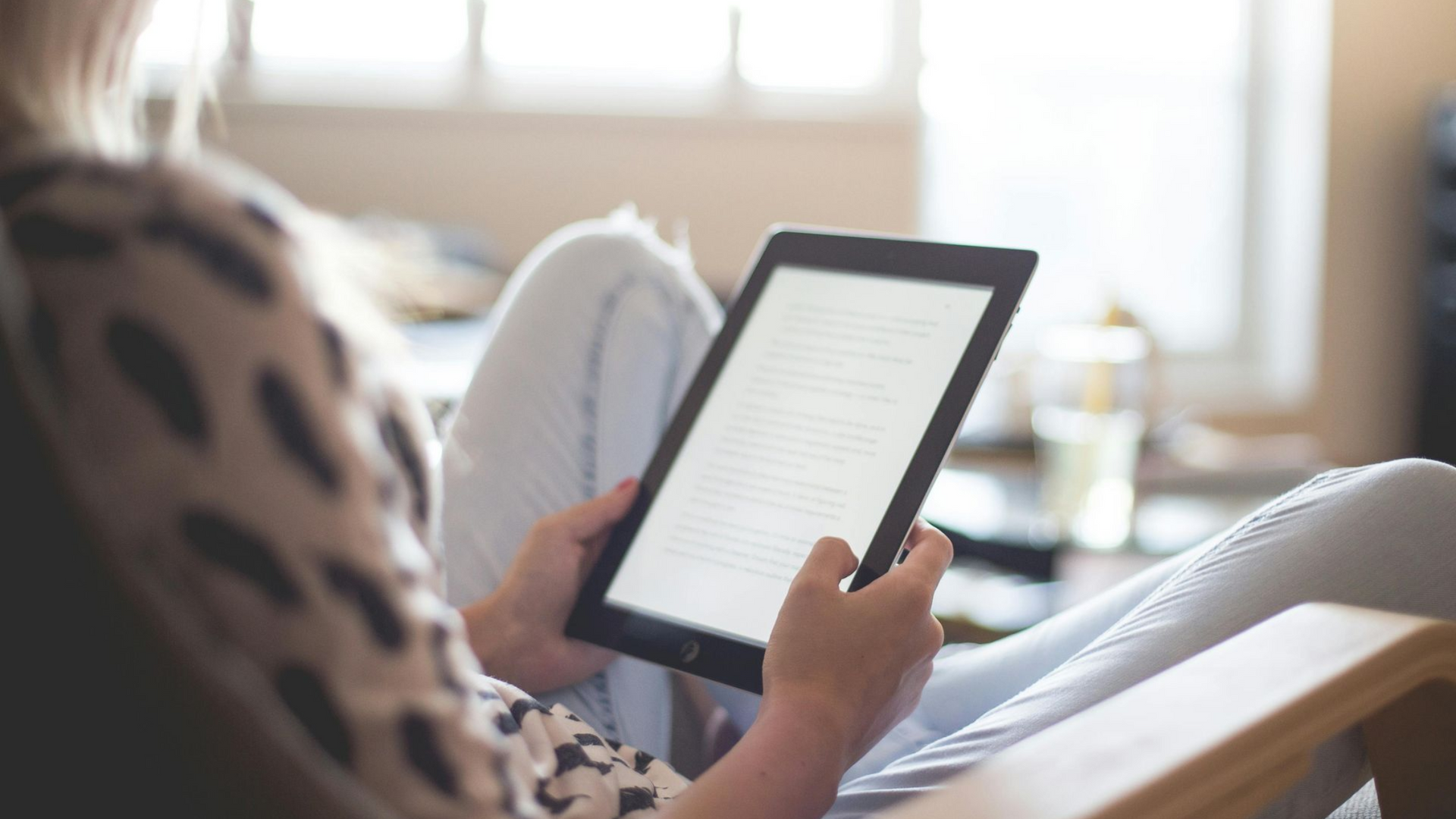 Person in a chair reading an e-reader; bright window in background.