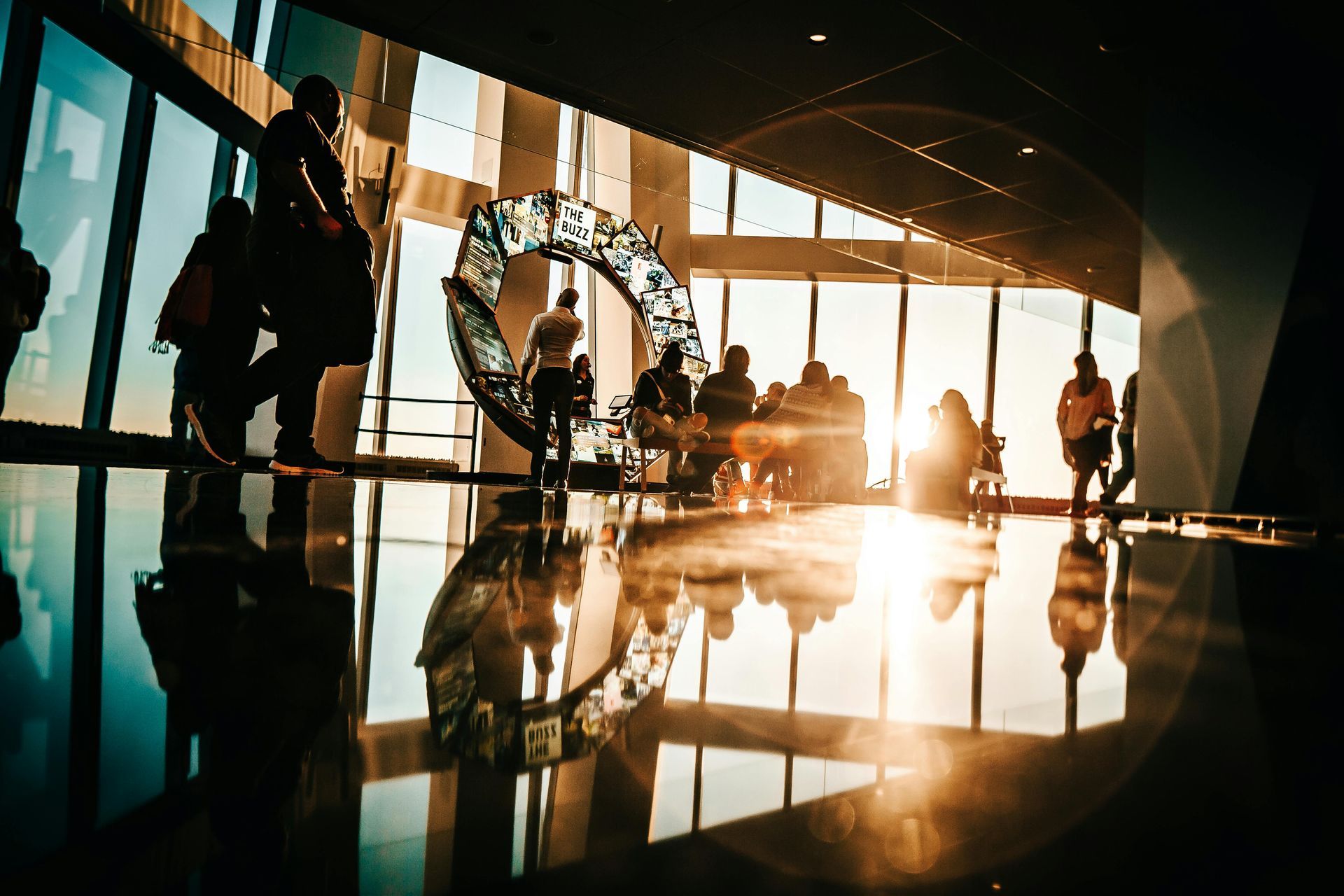 People in a modern building with large windows overlooking a bright sunset; artwork and reflections.