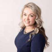 Woman with blonde hair wearing a navy blue dress smiles, posing against a white background.