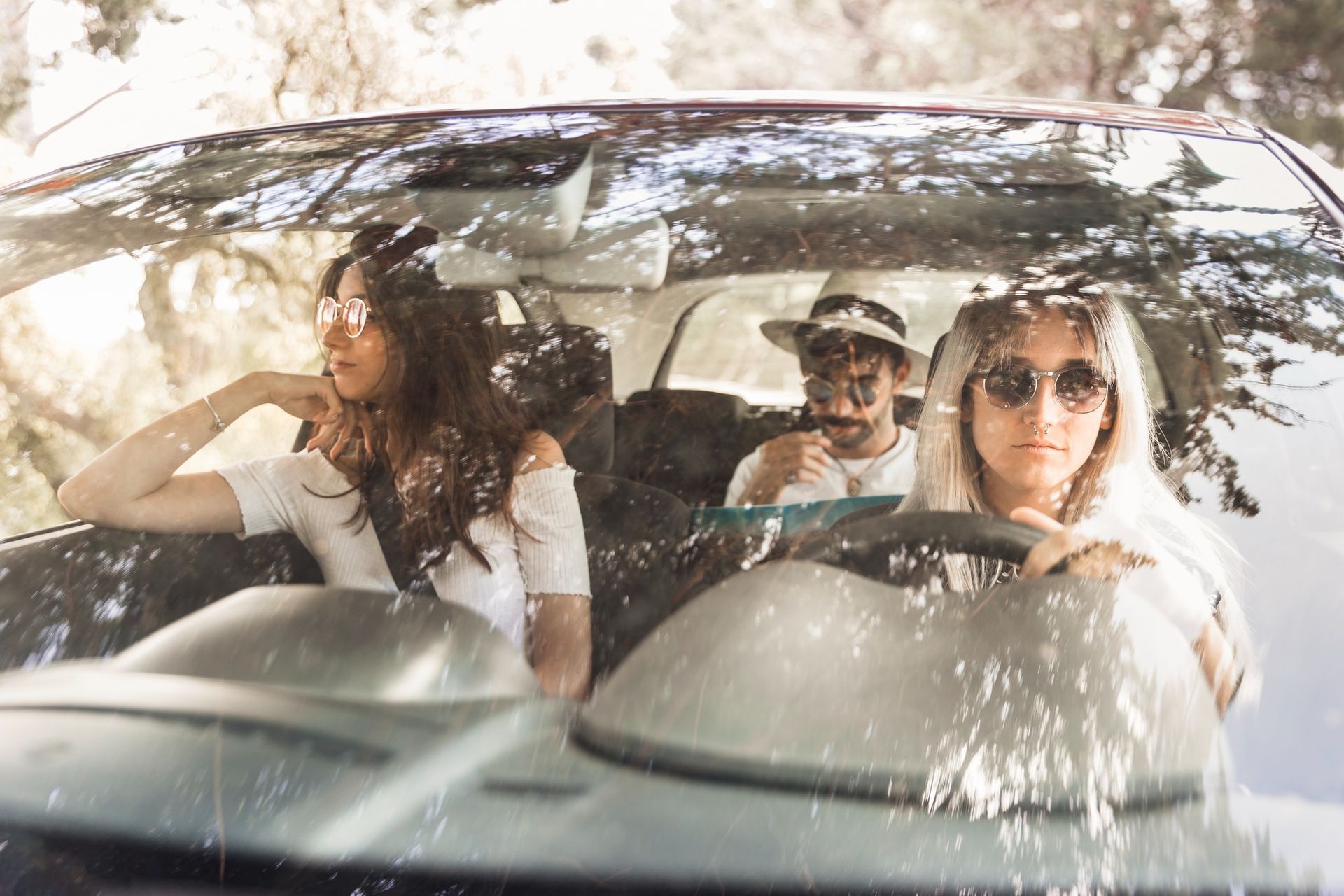 Three people ride in a car, with sunlight and tree shadows across the windshield.