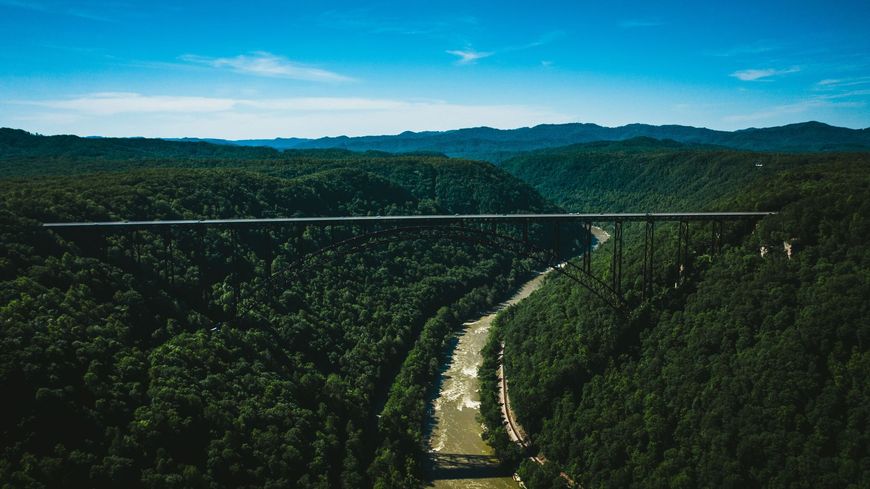 A high bridge spans a lush green gorge with a river below and forested hills in the distance.