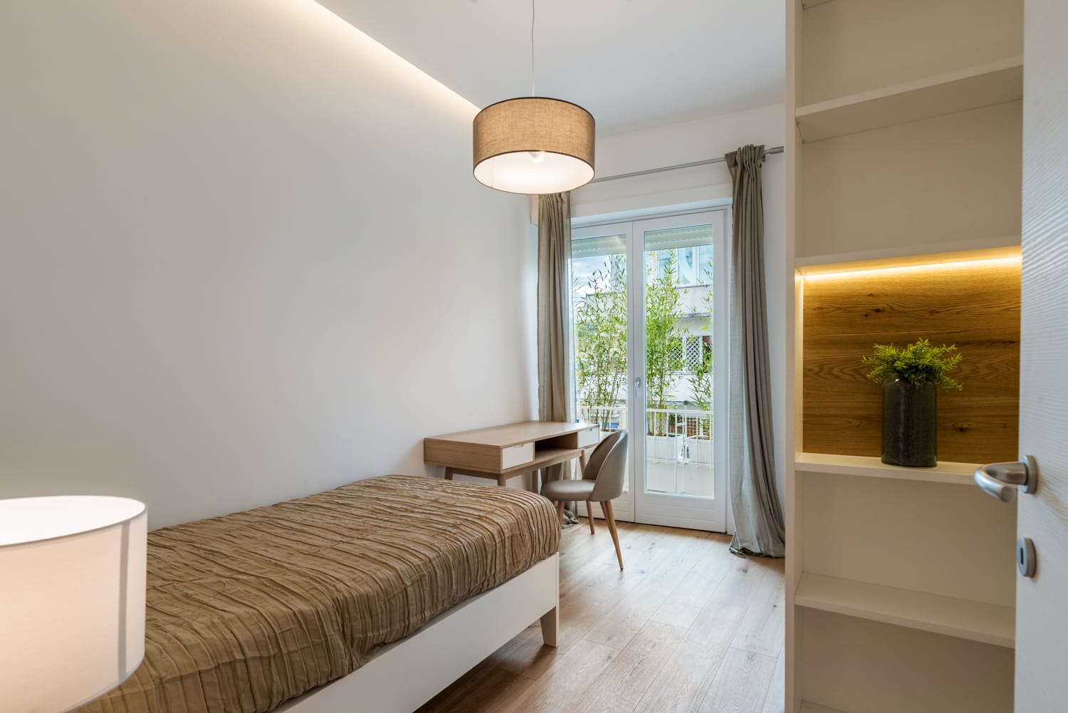Camera da letto luminosa e minimalista con letto singolo, scrivania accanto a porte finestre e illuminazione a incasso a caldo.
