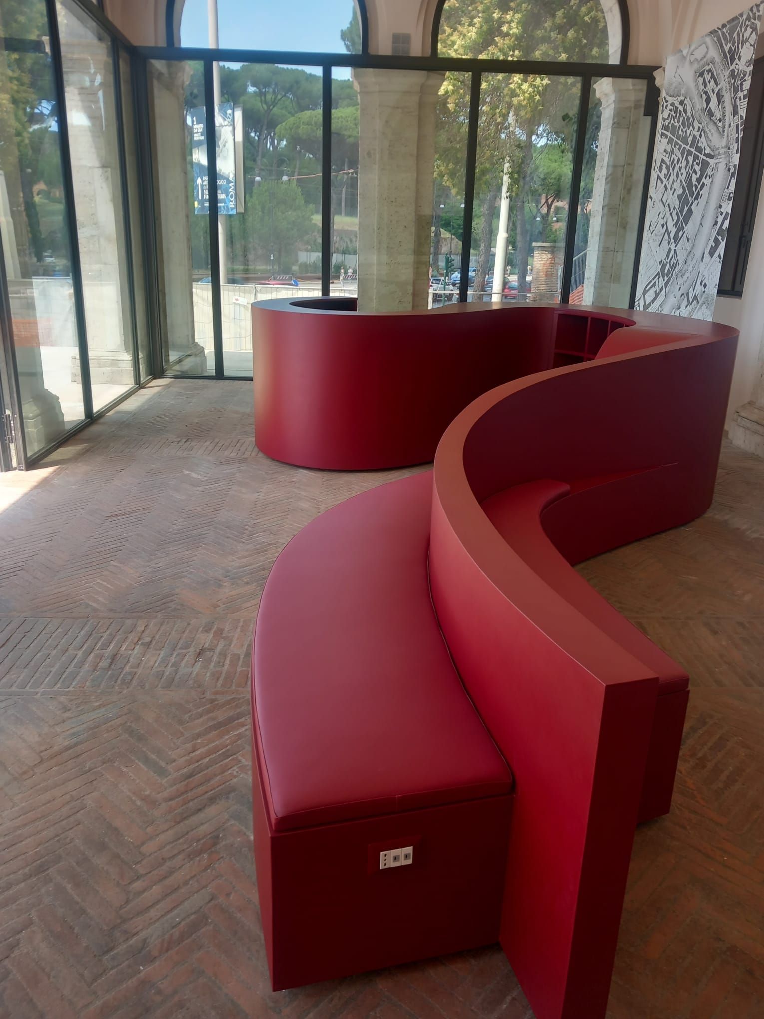Panchine curve rosse in una hall con pareti in vetro, pavimento in cemento e alberi all'esterno.