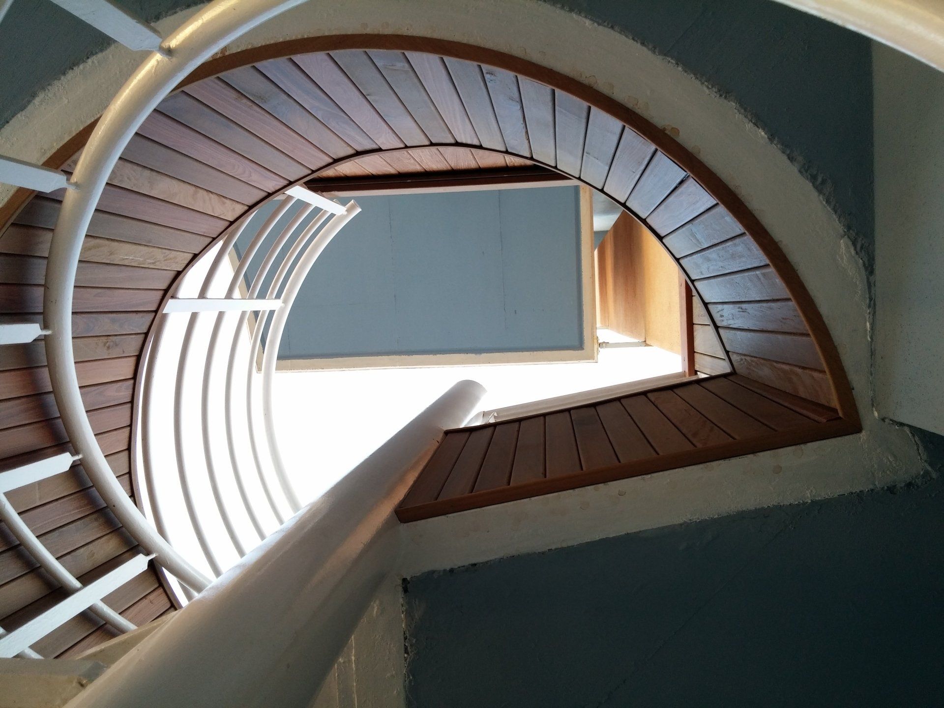 Scala a chiocciola vista dal basso, con ringhiere bianche e gradini in legno attorno a una luminosa apertura centrale.