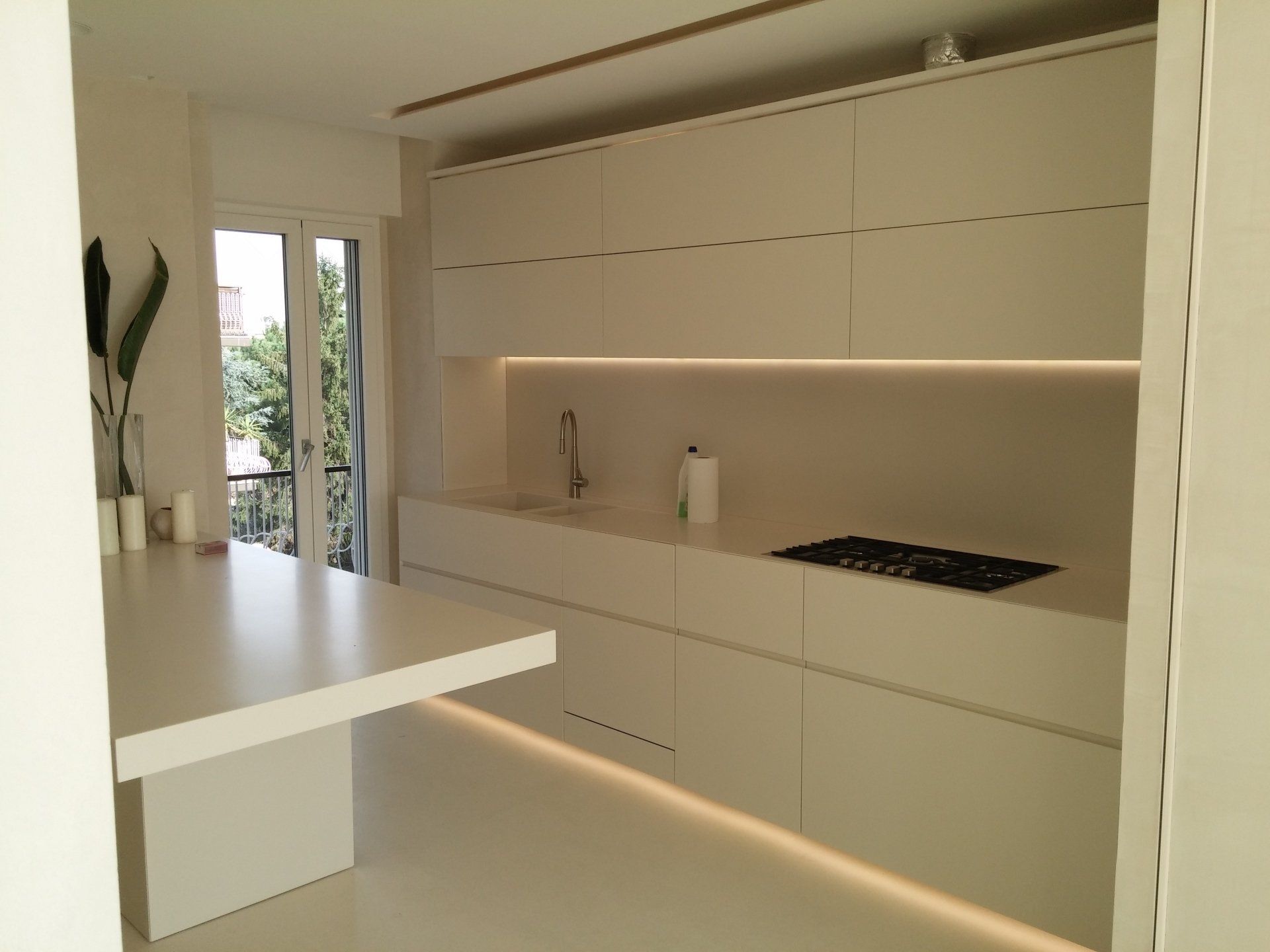 Cucina minimalista bianca con armadi a muro, isola centrale e illuminazione sottopensile a luce calda.