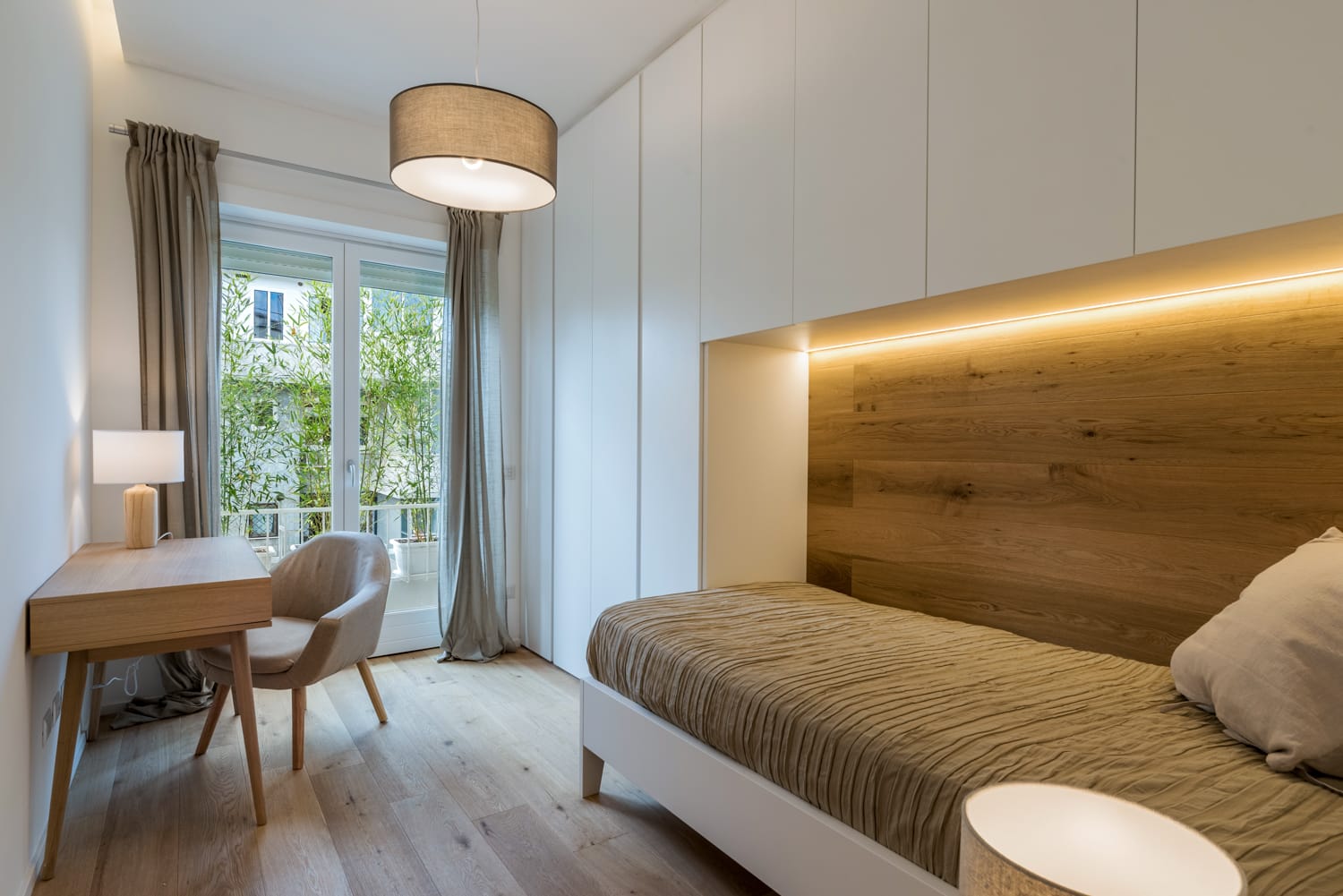Luminosa camera da letto moderna con letto singolo, parete con rivestimento in legno, scrivania e ampie porte finestre con tende.
