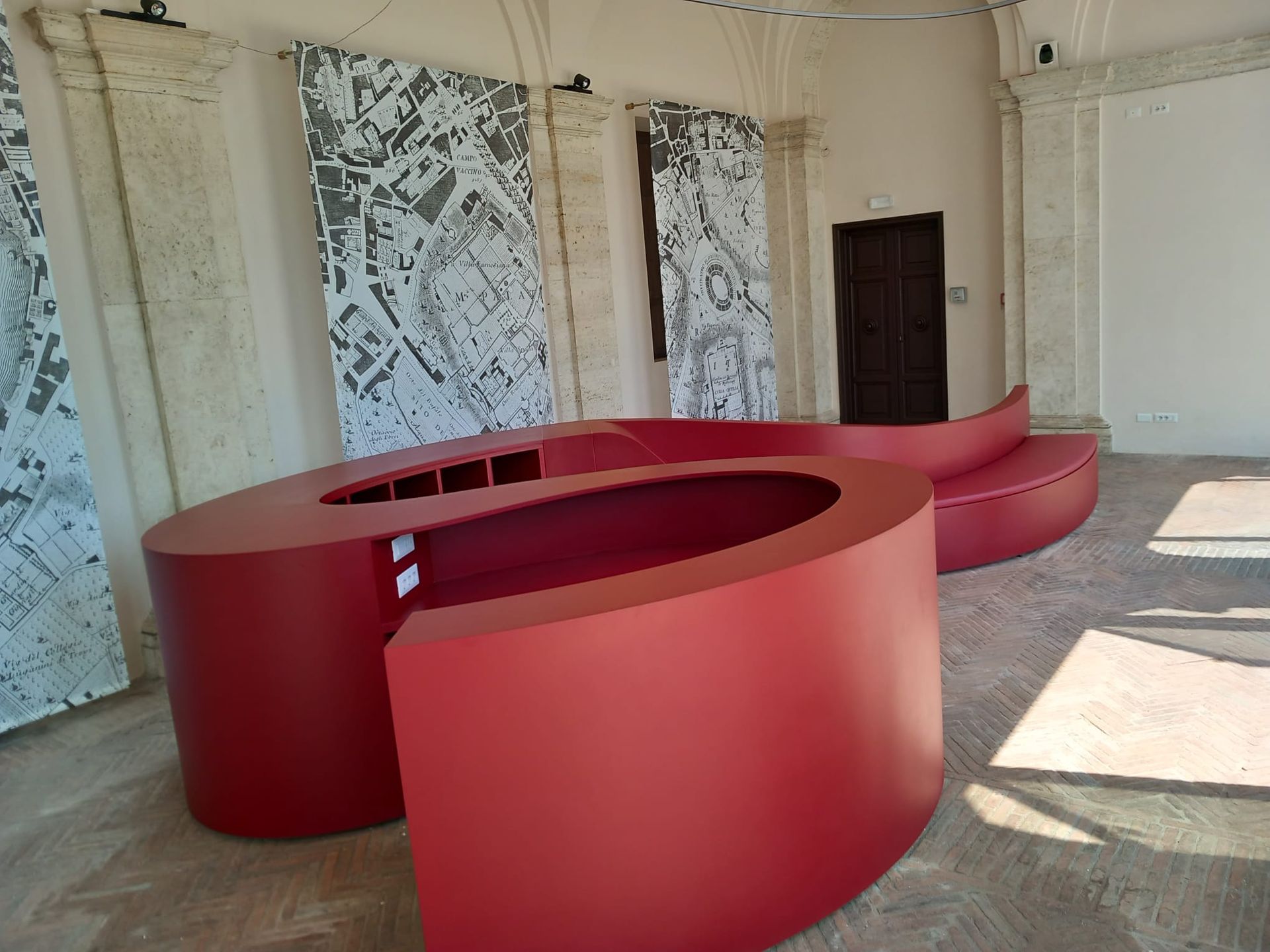Banconi reception rossi e curvi in ​​una stanza spoglia e soleggiata con pareti consumate e finestre sbarrate.