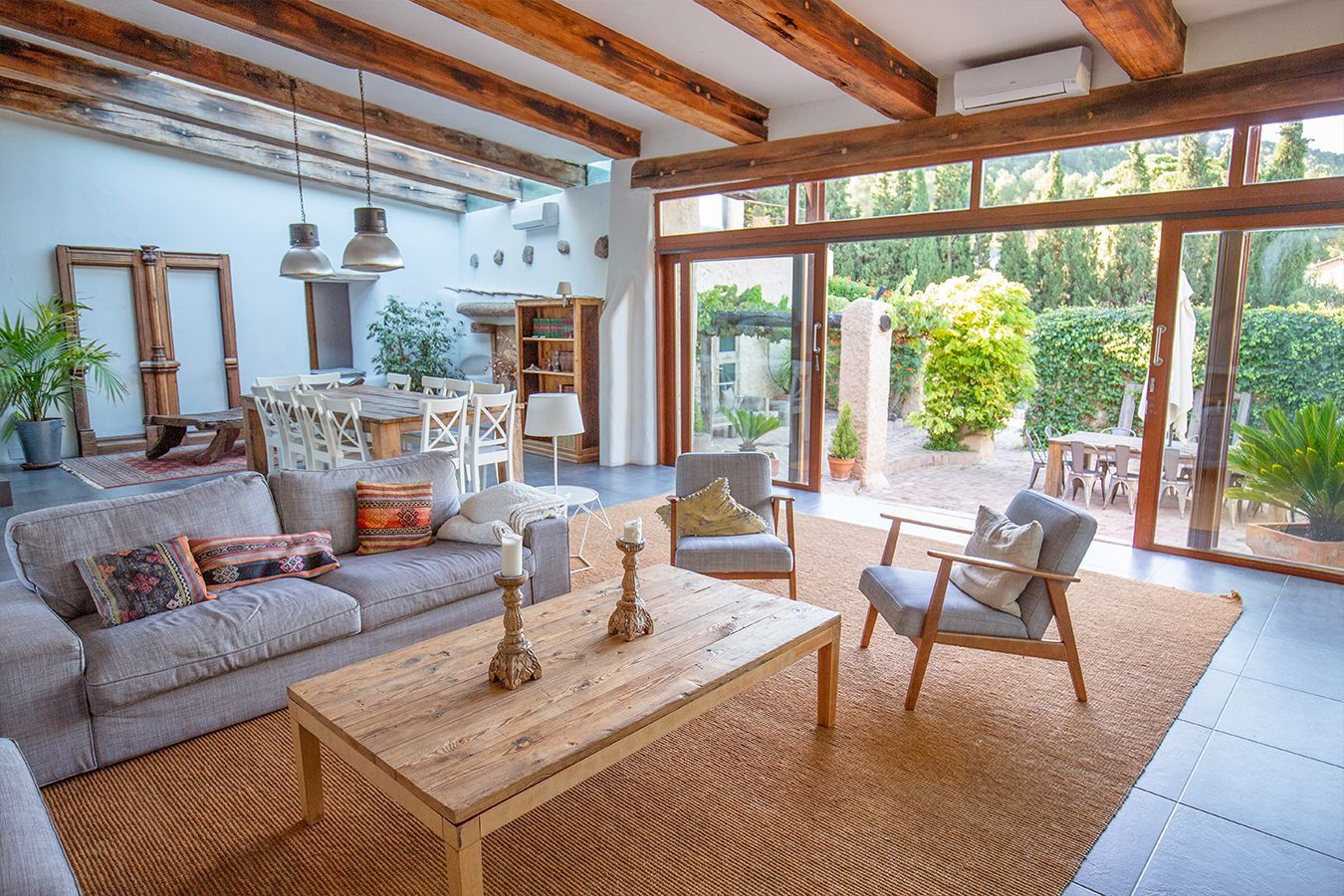 Luminoso soggiorno con divano grigio, travi in legno, tappeto intrecciato e porte aperte che conducono a un patio soleggiato.