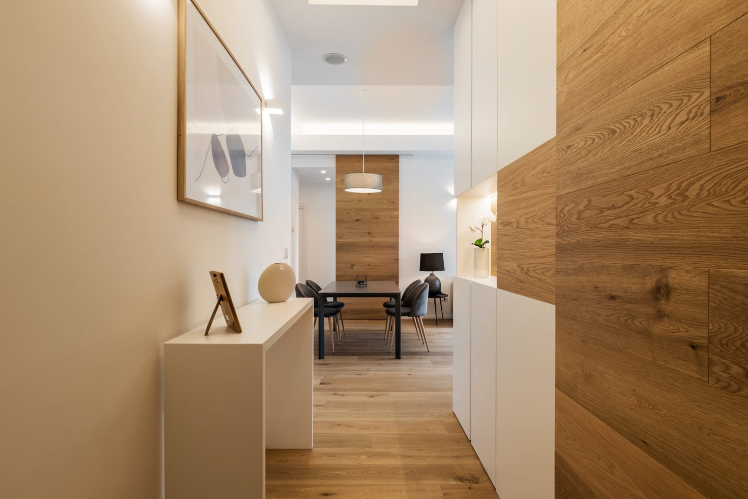 Corridoio moderno con pavimento in legno, mobili bianchi e zona pranzo in fondo.