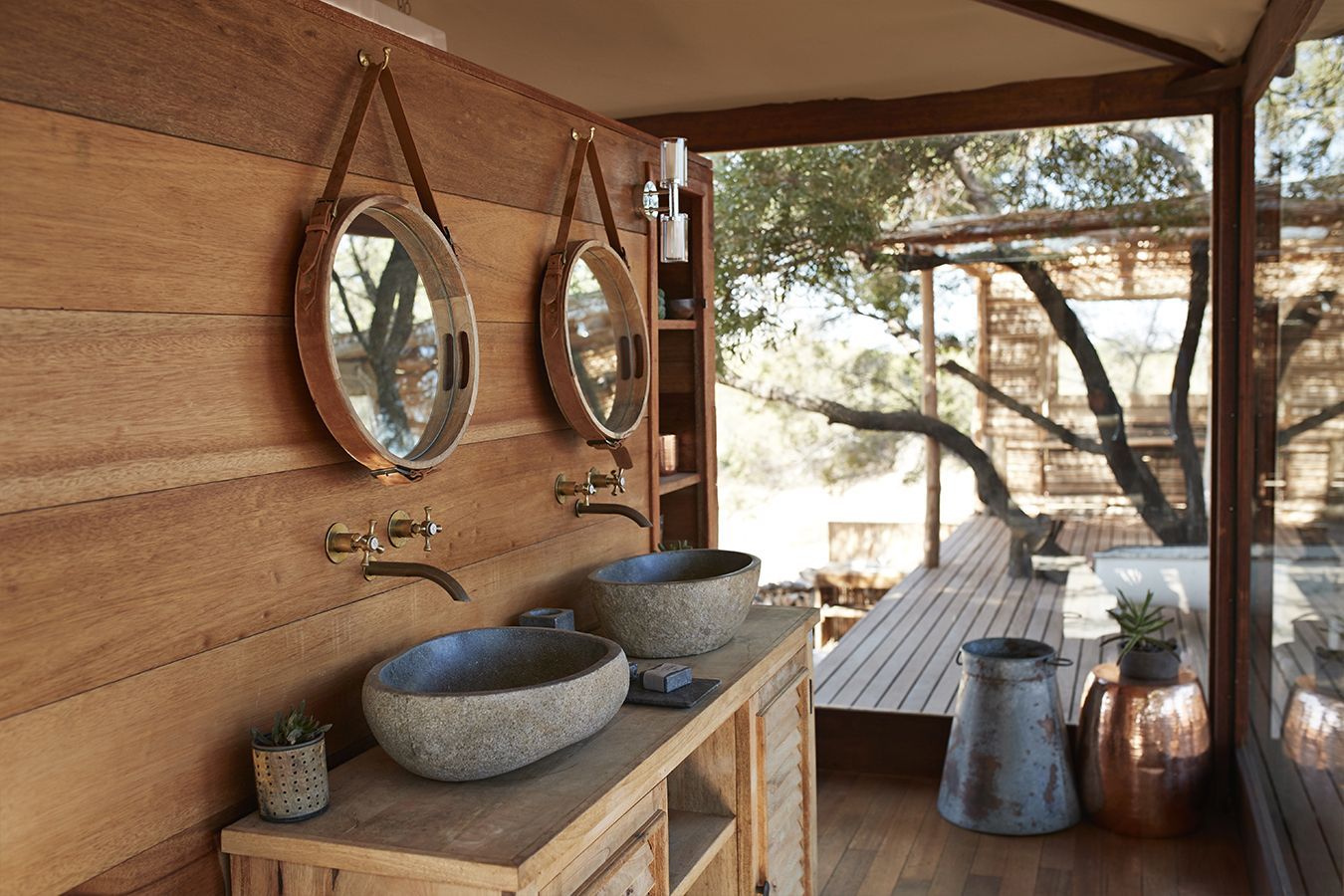 Zona bagno esterna rustica con due specchi, lavandini in pietra e terrazza in legno con vista sugli alberi.