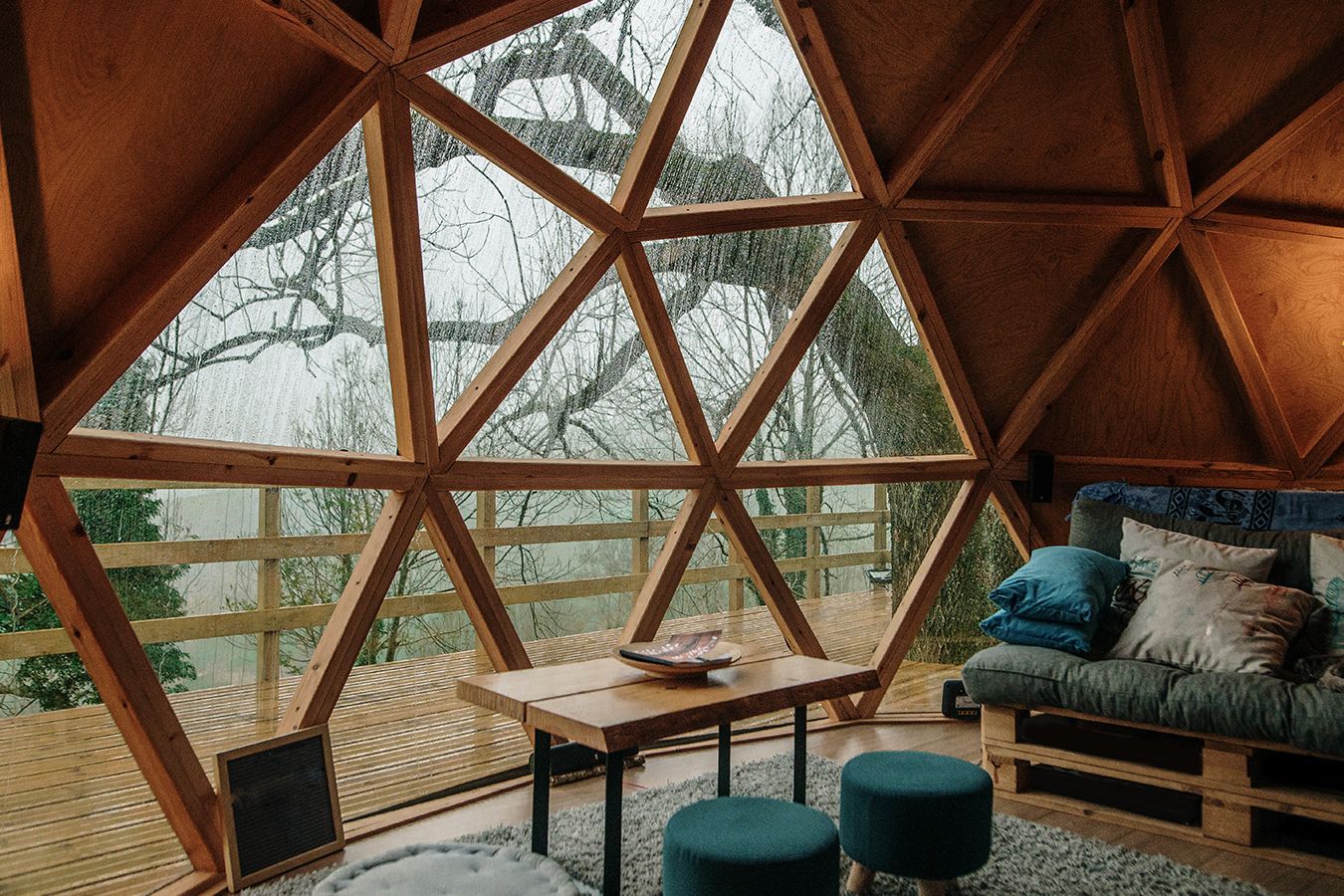 Interno a cupola geometrica in legno con divani, tavolo, sgabelli e vista sulla foresta attraverso ampie finestre triangolari.