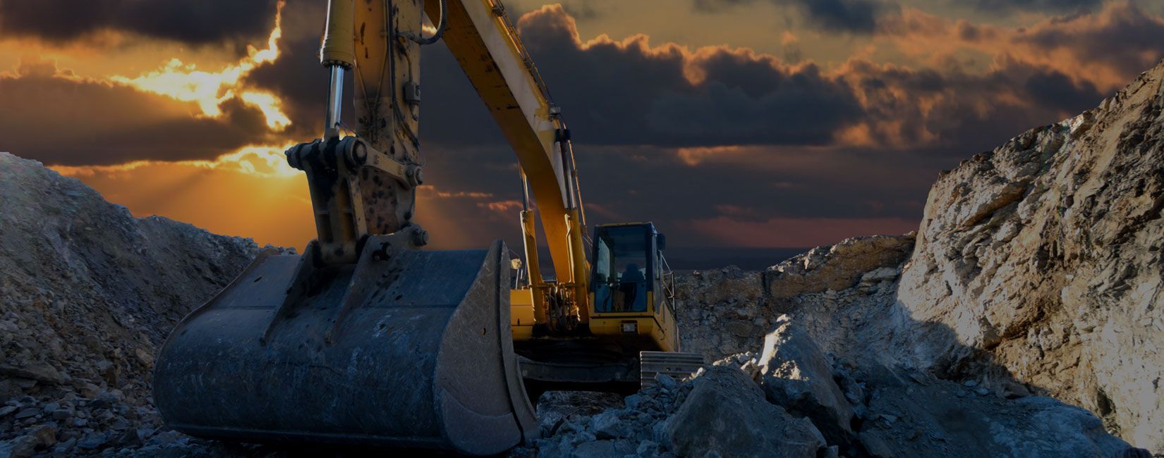 A yellow excavator is digging a hole in a rocky area at sunset.