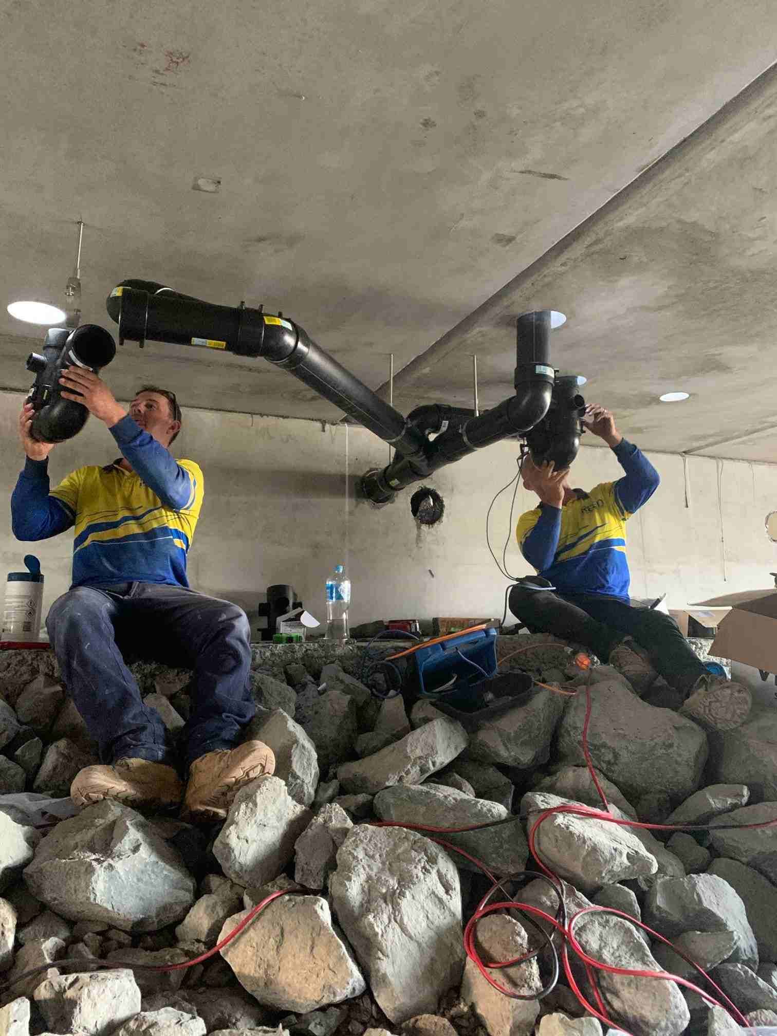 Two Men Are Sitting on a Pile of Rocks Working on Pipes — Reed Plumbing Industries in Mackay, QLD