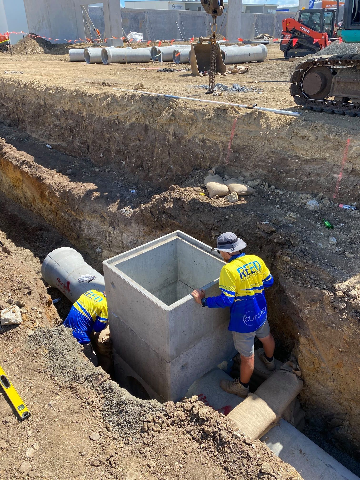 Two Construction Workers Are Standing in a Trench Looking at a Pipe Being Installed In The Ground — Reed Plumbing Industries in Mackay QLD