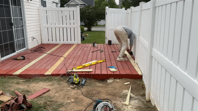 Man building a deck; using saw against a white fence. Red and light-colored wood on the deck. Outdoors, sunny.