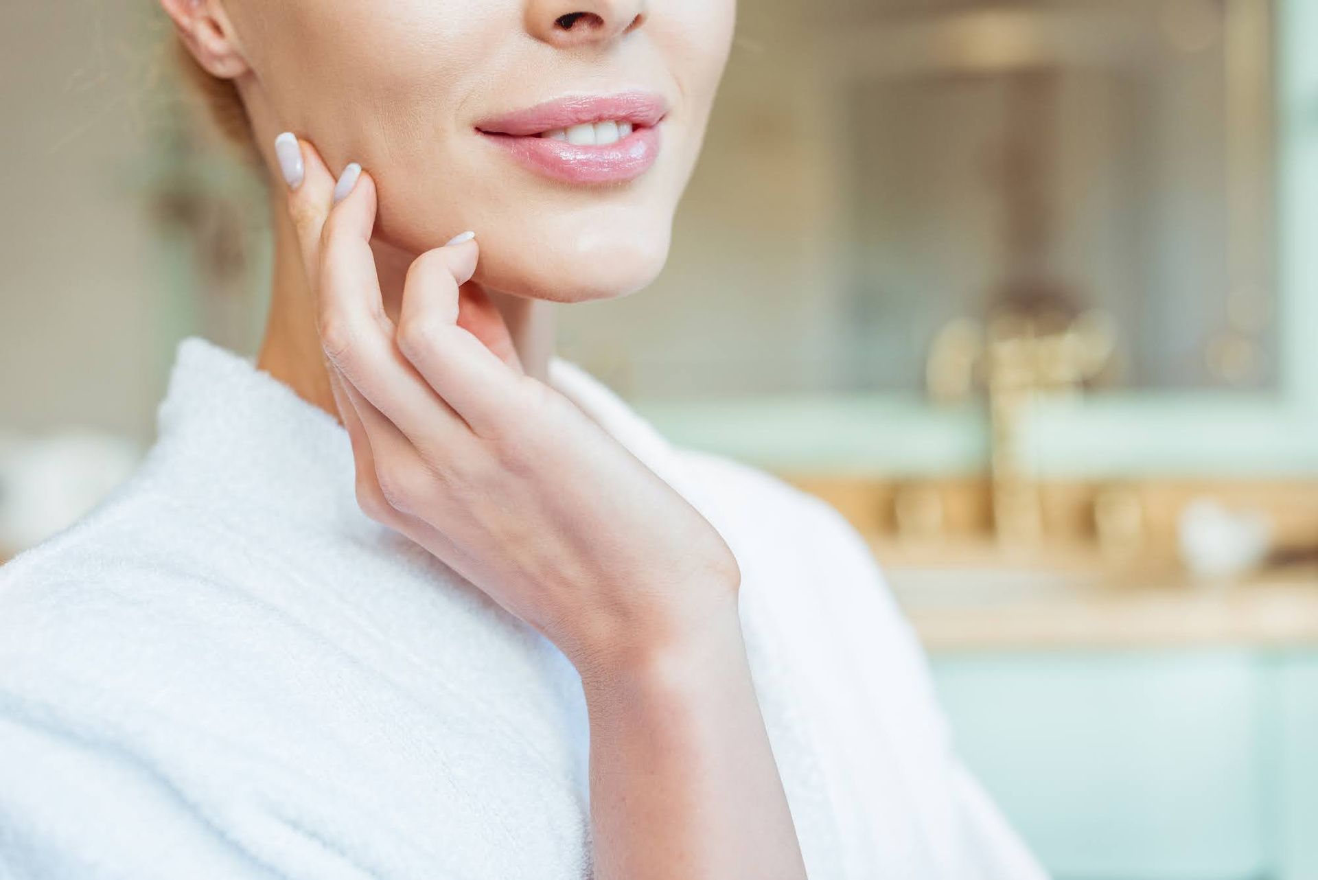Woman in a white robe gently touching her cheek, smiling slightly, in a bathroom setting.
