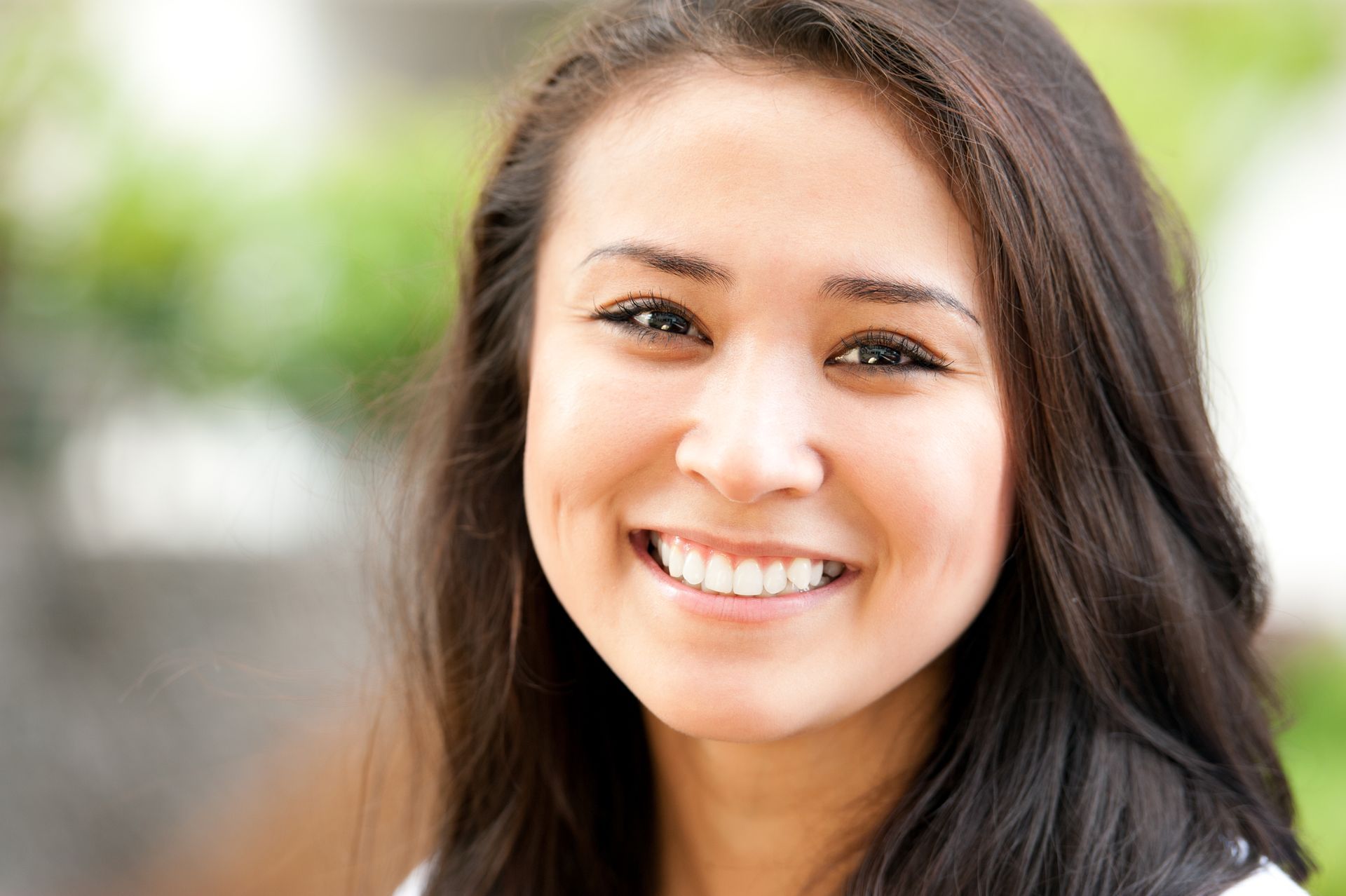 Smiling woman with dark hair, looking at the camera, outdoors with blurred background.