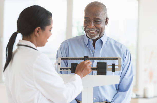 Doctor adjusting scale for patient. Smiling person in blue shirt stands on a scale in a bright room.