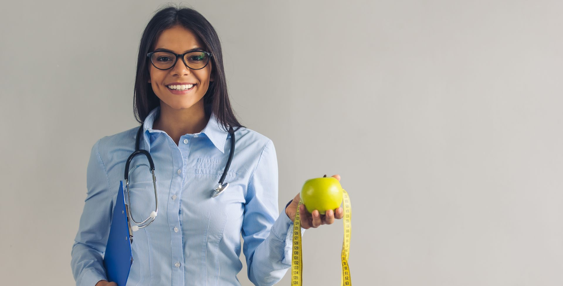 Woman in glasses, holding apple and measuring tape, smiling, wearing stethoscope.