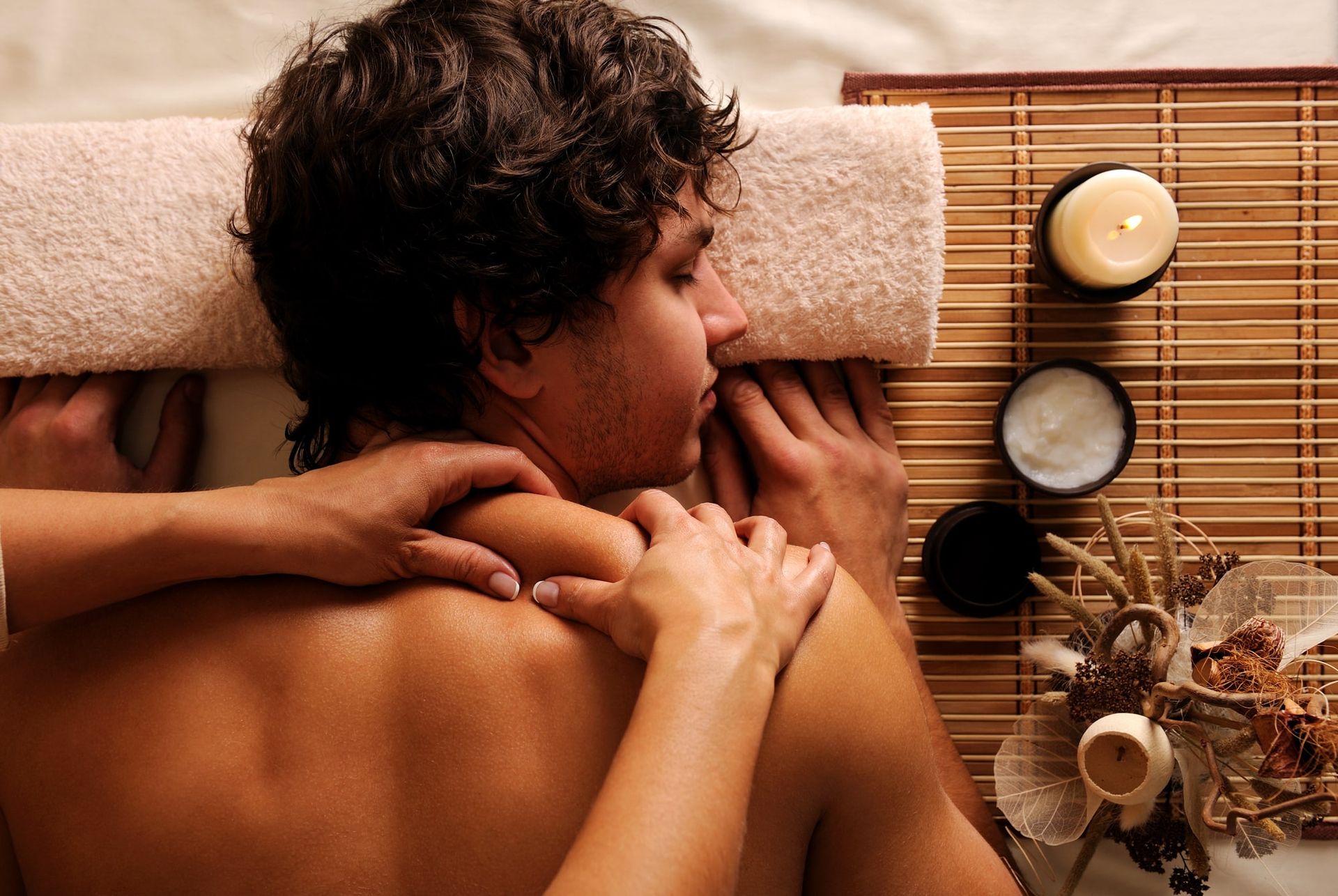 Man receiving a shoulder massage at a spa; lit candle and cream jar on a bamboo mat.