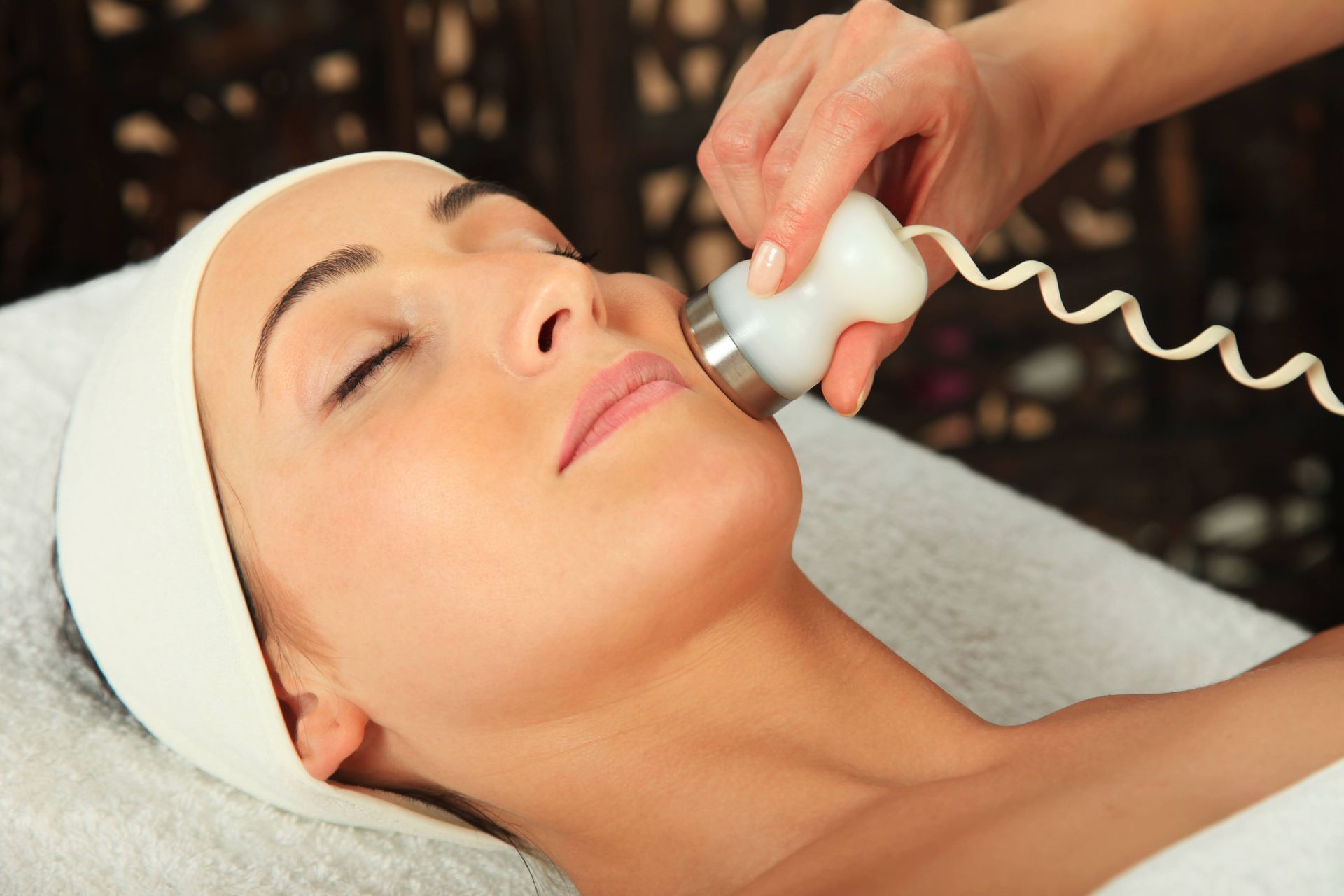 Woman receiving facial treatment with a handheld device on a spa table, eyes closed.