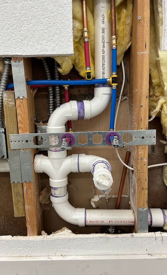 Exposed plumbing in a wall. White PVC pipes with metal support bracket. Copper and blue PEX water lines.