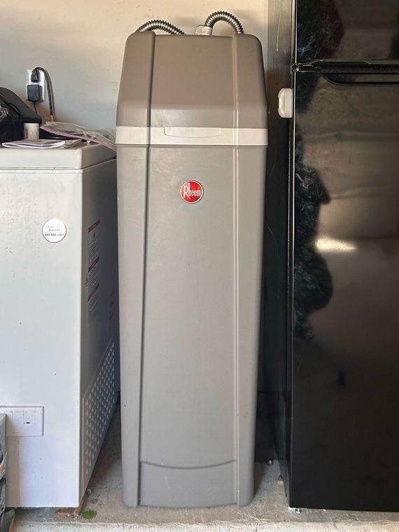 Gray Rheem water softener next to a white appliance and black refrigerator in a utility area.