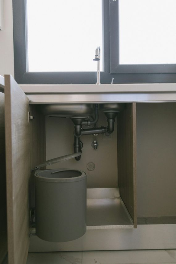 Kitchen cabinet under sink with plumbing, trash can, and open door. Window in the background.