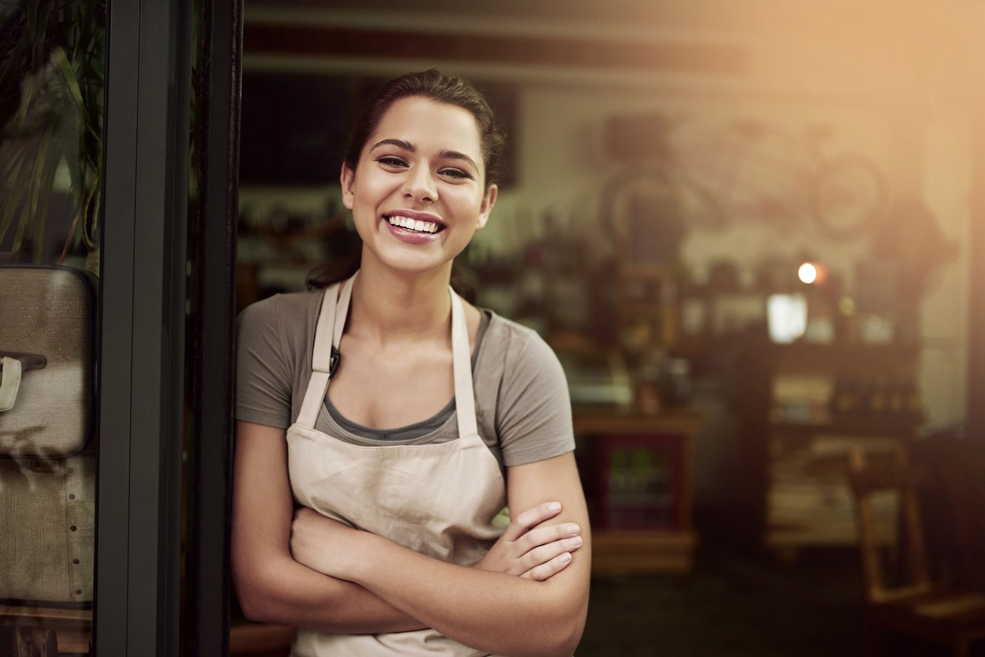 A woman in an apron is standing in the doorway of a restaurant with her arms crossed and smiling.