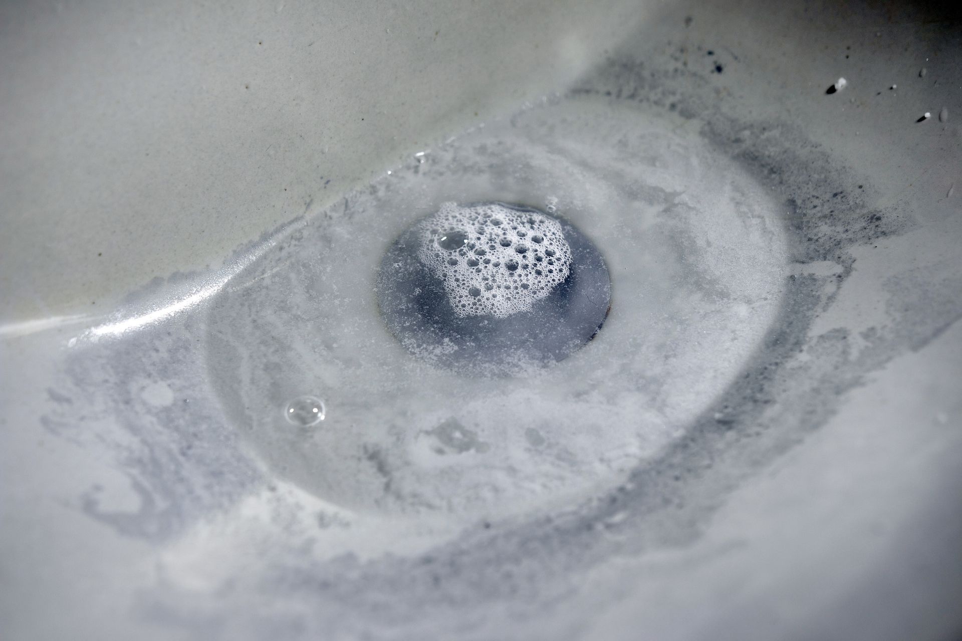 A close-up view of a white bathroom sink drain filled with gray, soapy water and bubbles.