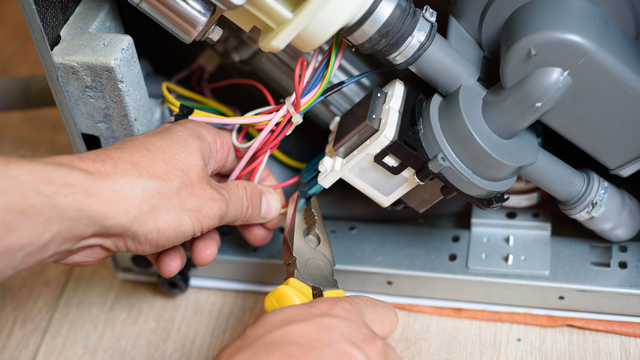 A person is fixing a dishwasher with a pair of pliers.