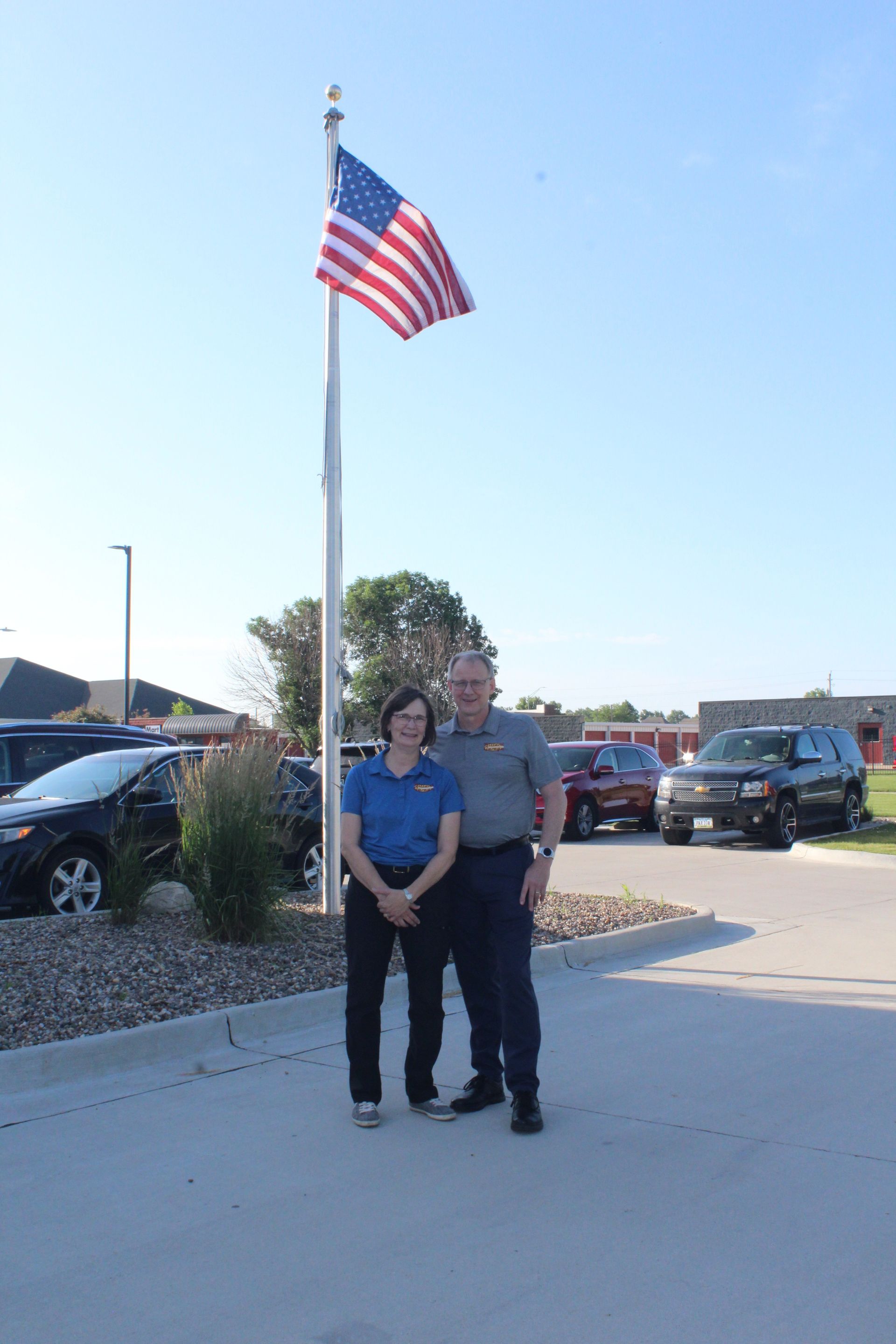 Two people standing on a sidewalk near a parking lot with a large American flag overhead