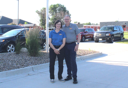 Two people standing together on a sunny sidewalk in a parking lot, with parked cars and landscaping behind them.