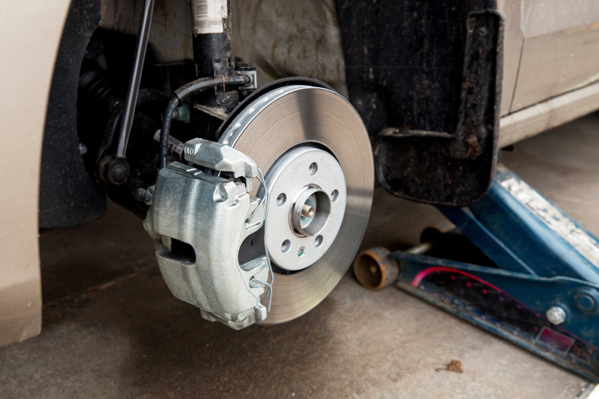Car’s front brake disc and caliper visible with wheel removed, blue jack on the garage floor