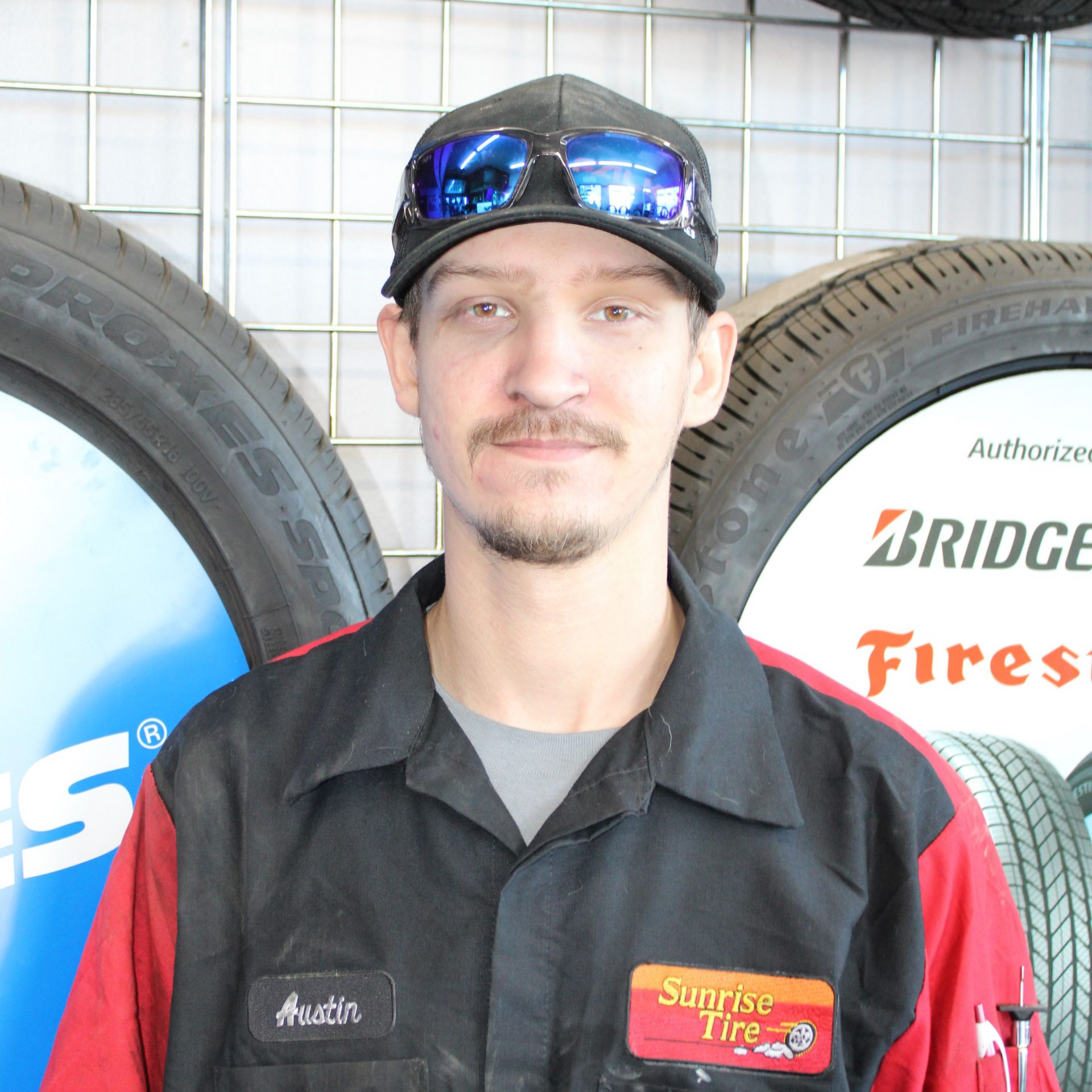 Mechanic in a black shirt and cap standing between large tires in a tire shop, with glasses on his cap.