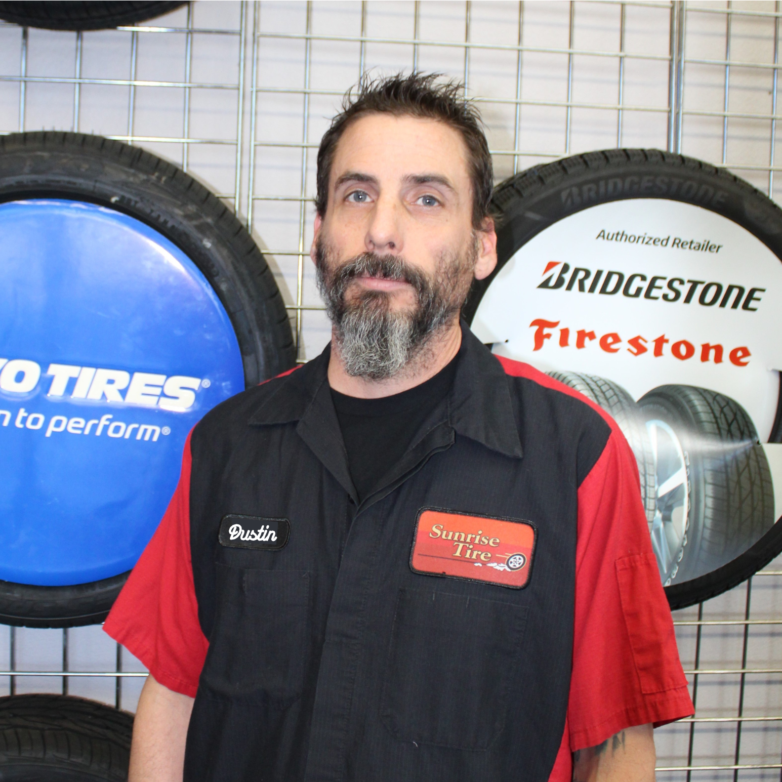 Man in a black and red work shirt standing in front of tire display signs with Bridgestone and Firestone logos
