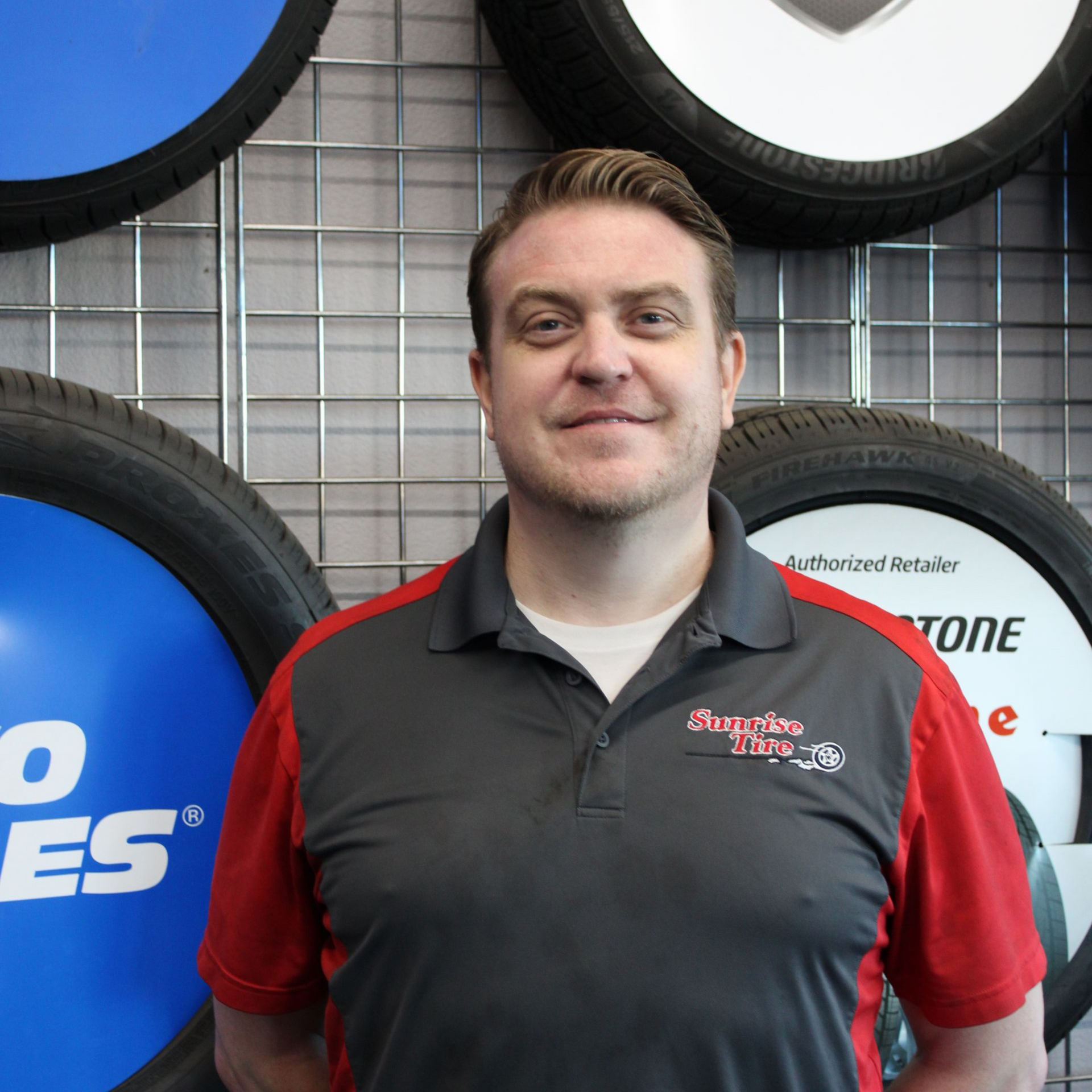 Man in gray and red shirt, smiling, standing in front of tires.