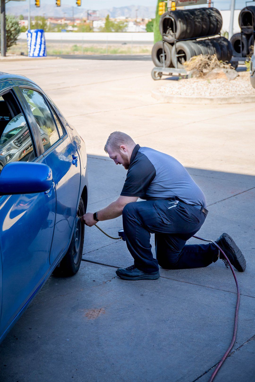 Mechanic kneels beside a blue car, inspecting the wheel area with a tool in an outdoor lot