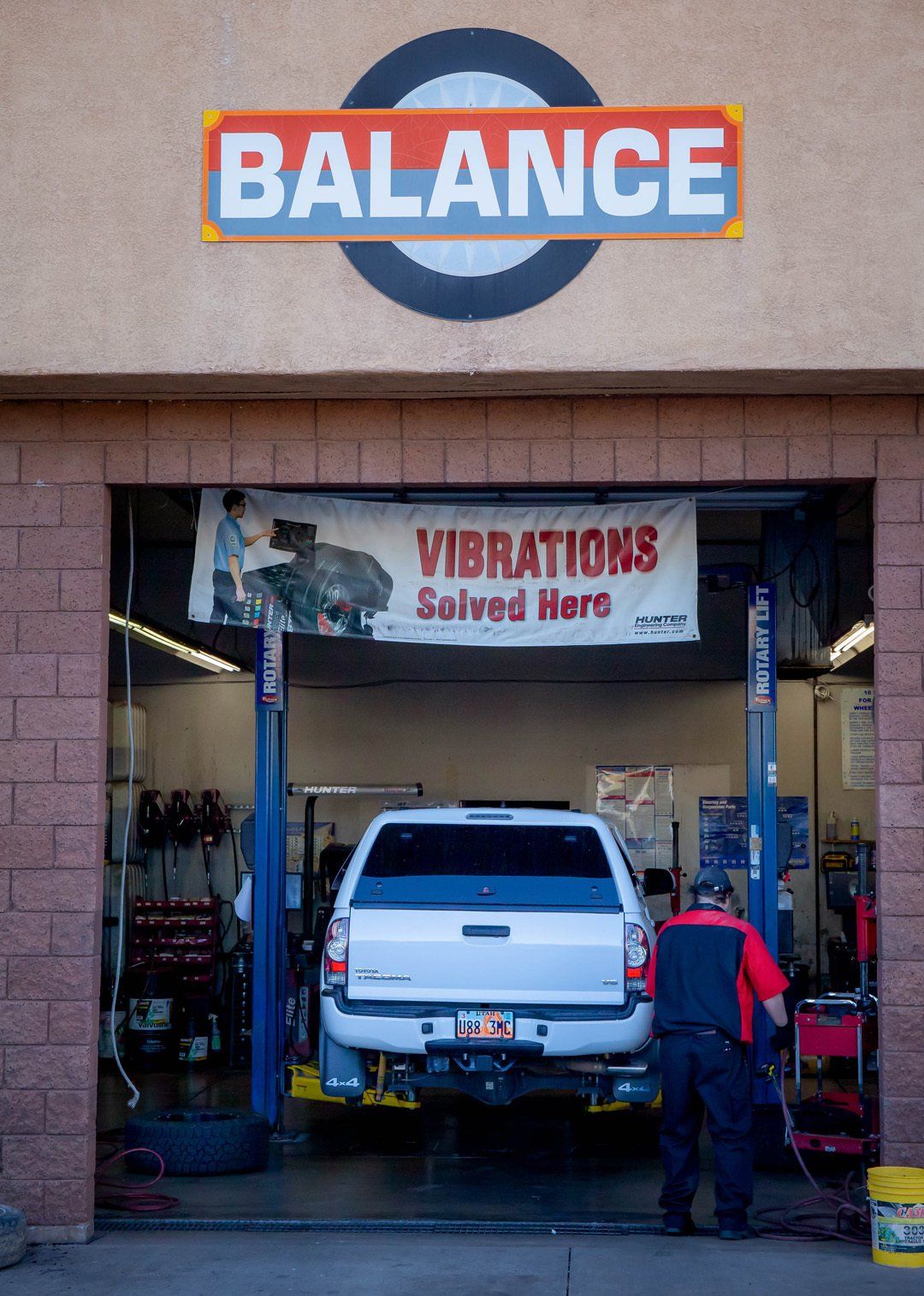 Auto repair shop with a white SUV on a lift and a mechanic underneath a BALANCE sign