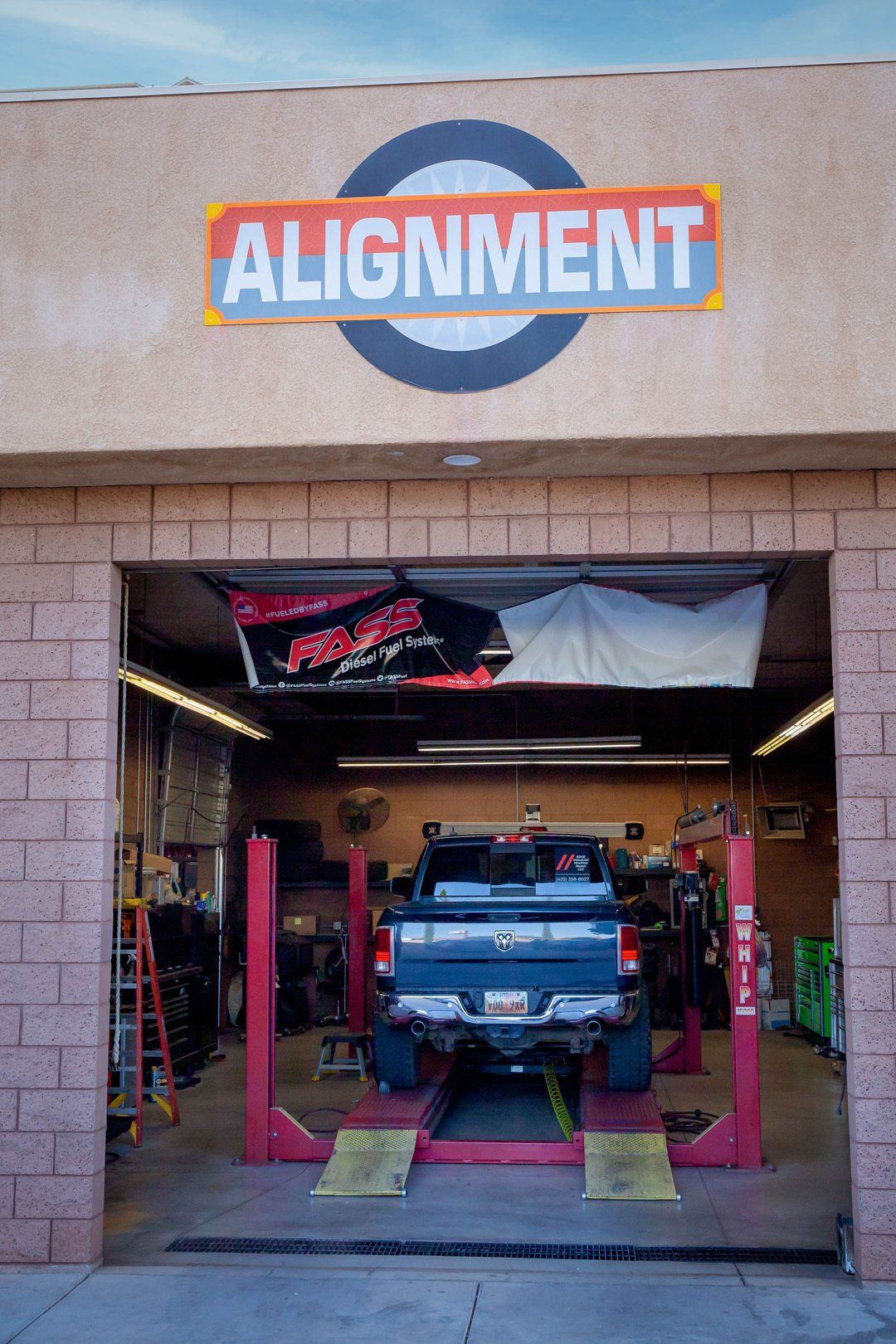Car in an auto shop bay beneath an “ALIGNMENT” sign, with a truck parked inside the service area.
