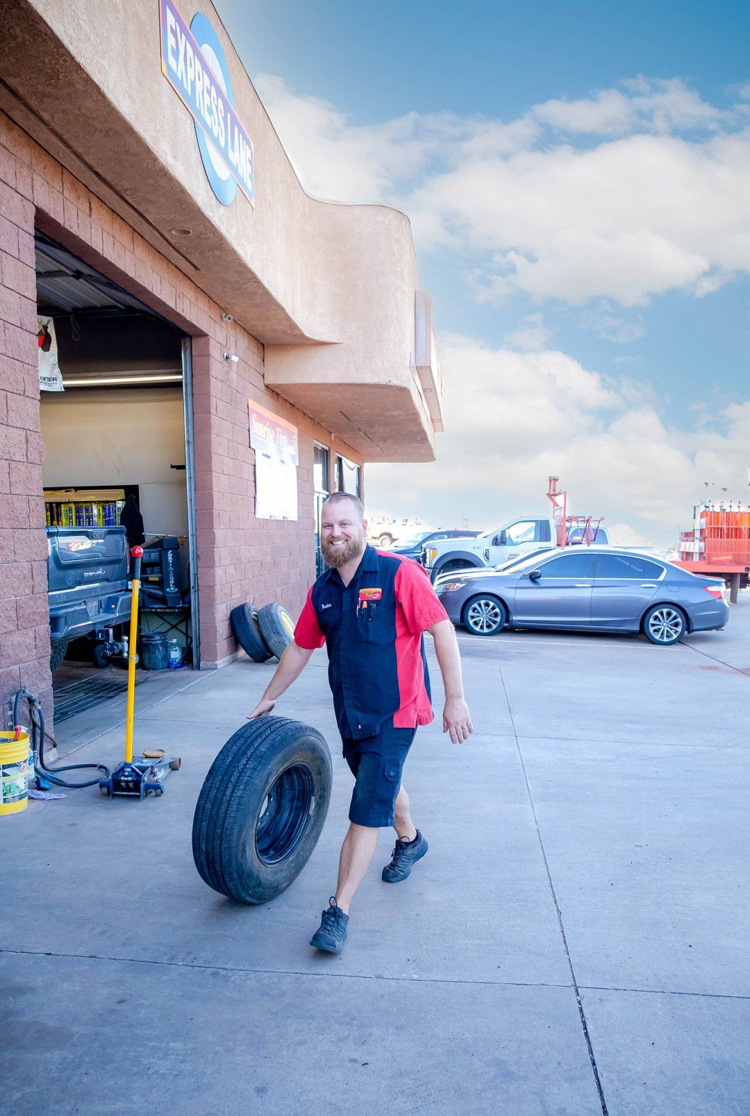 Man in red shirt walking a tire outside an auto shop on a sunny day