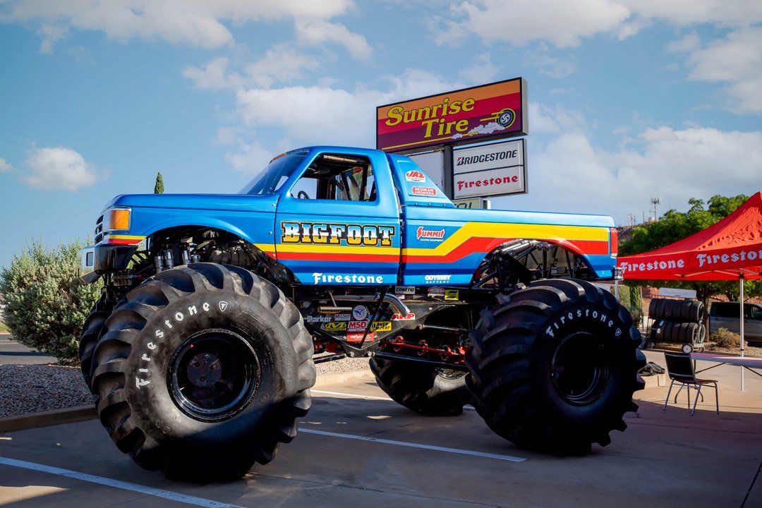 Blue monster truck with oversized tires at an outdoor event, parked beside a red canopy.
