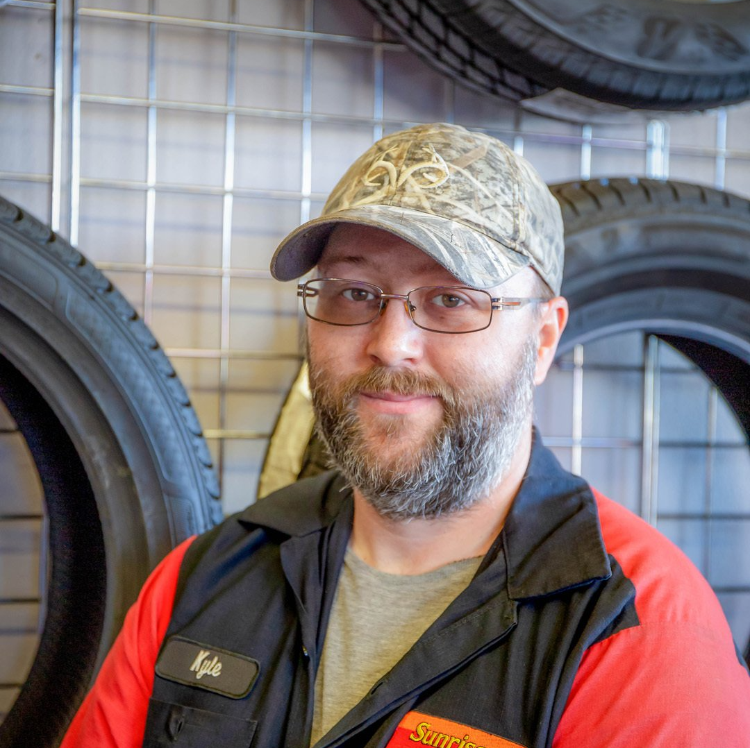 Man in camouflage cap and red jacket smiling in a tire shop, with stacked tires behind him