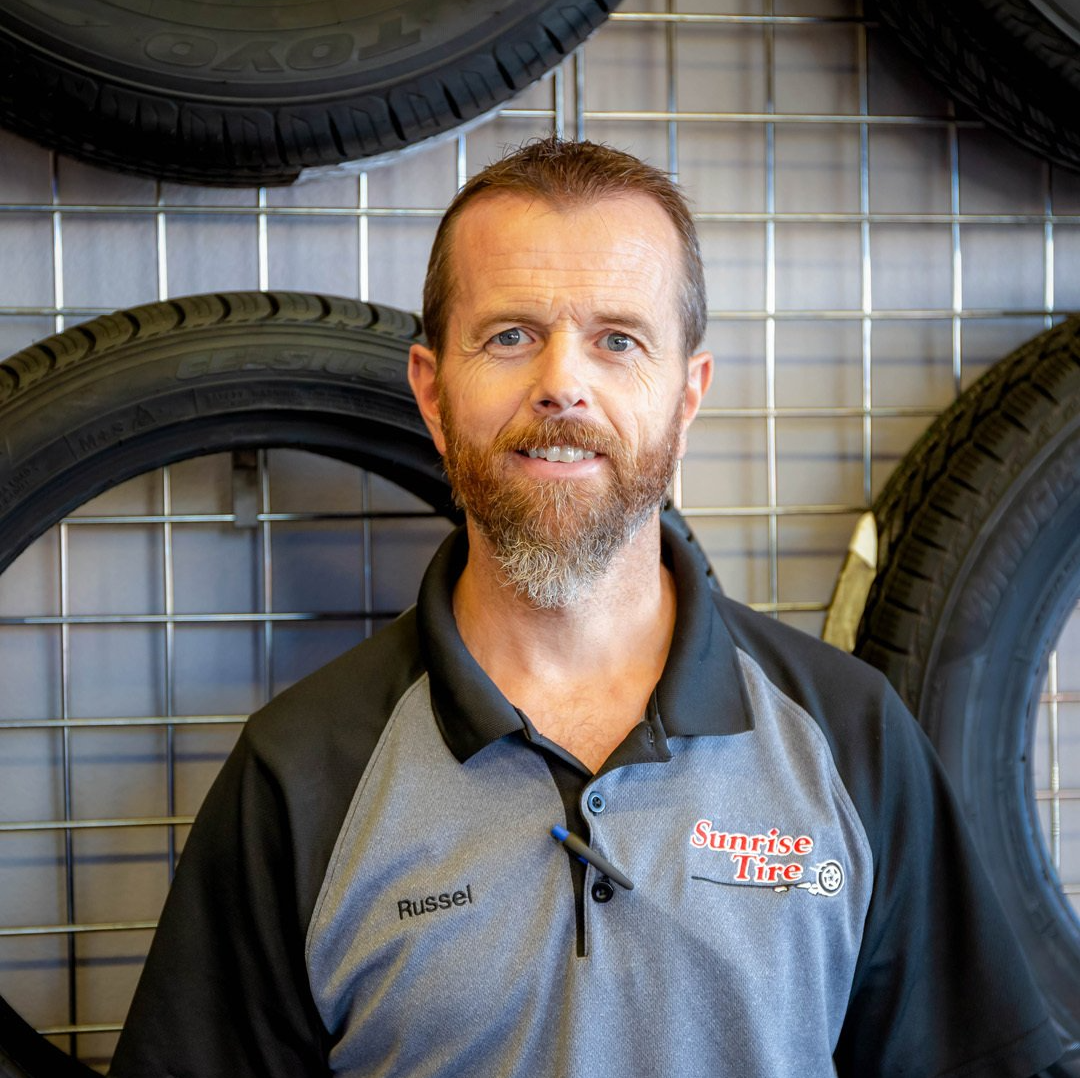 Man in a gray and black shop shirt standing before display tires on a metal grid wall