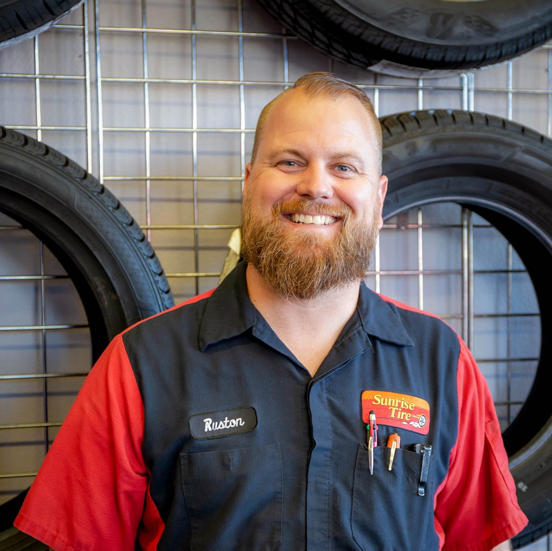 Smiling tire shop employee in red and navy uniform standing in front of stacked tires